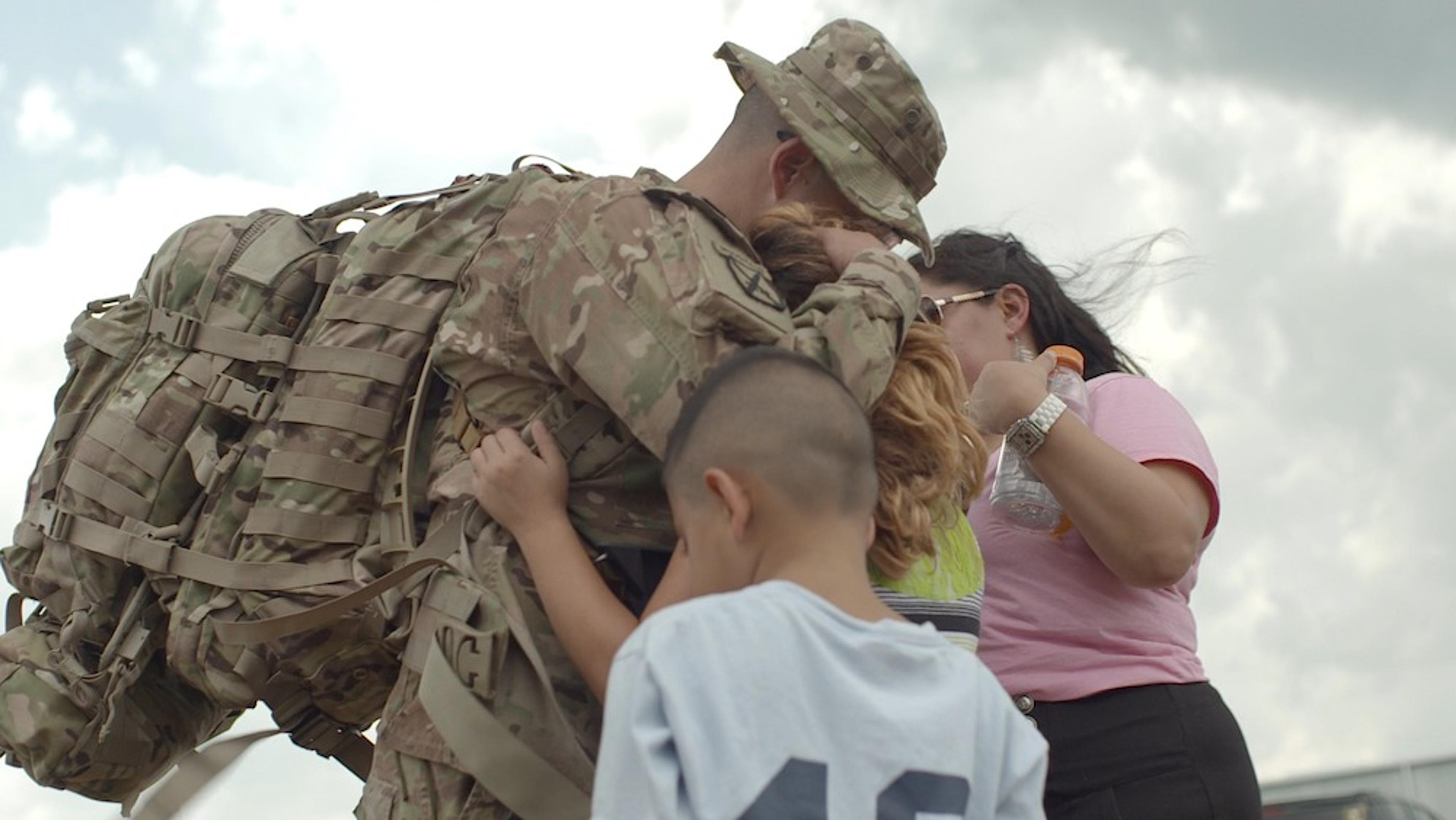 A soldier in camouflage gear hugging a family, including a woman and two children, against a cloudy sky.