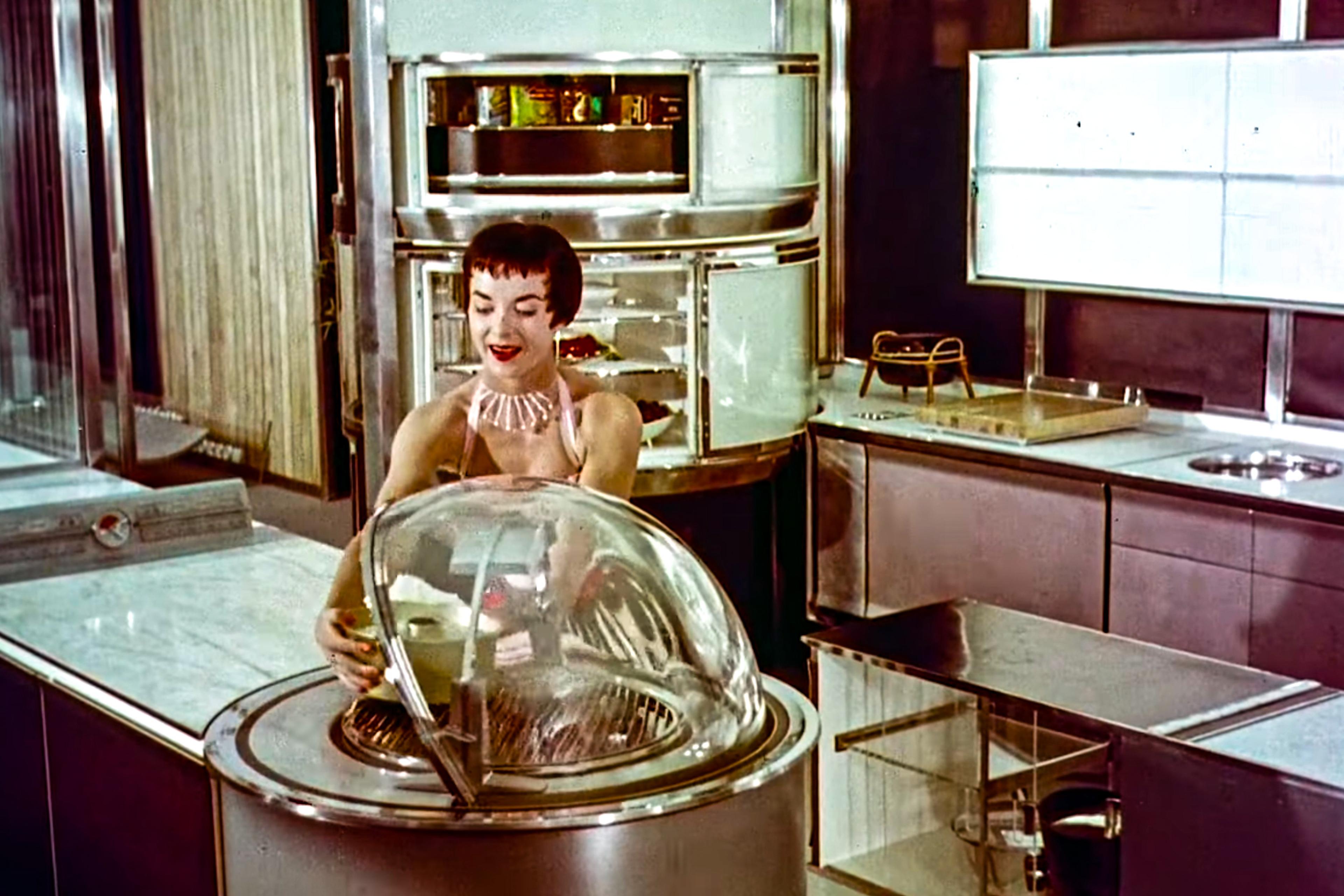 Vintage photo showing a woman in a retro futuristic kitchen operating a round appliance with a clear dome lid.