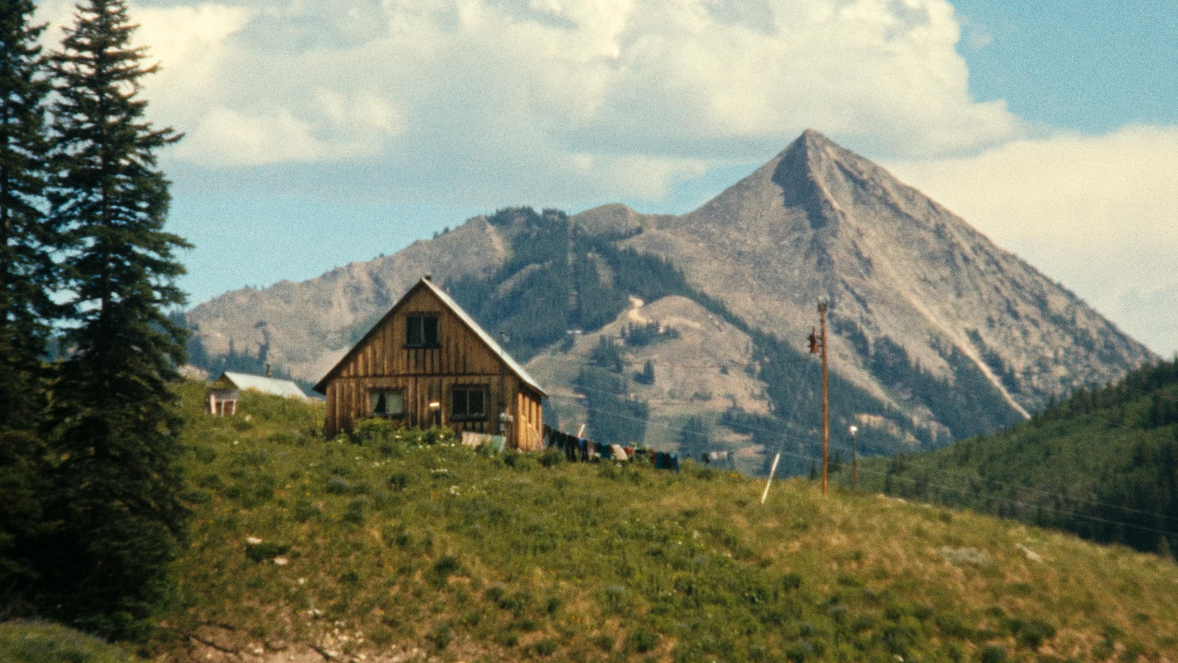 A wooden cabin on a grassy hill with a mountainous backdrop and cloudy sky in the distance.