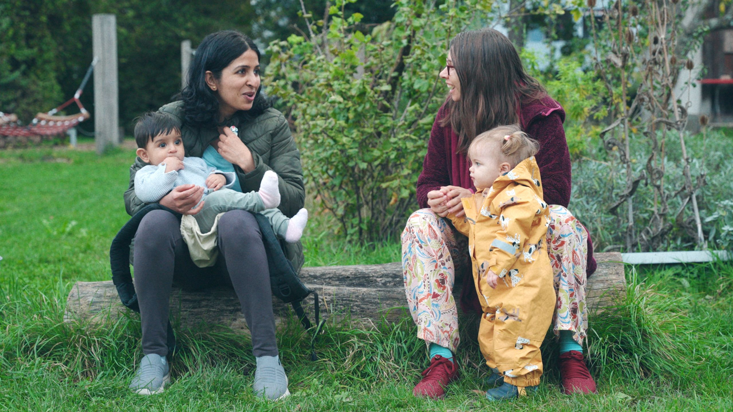 Two women sitting on a log outdoors, each holding a child, with greenery in the background.