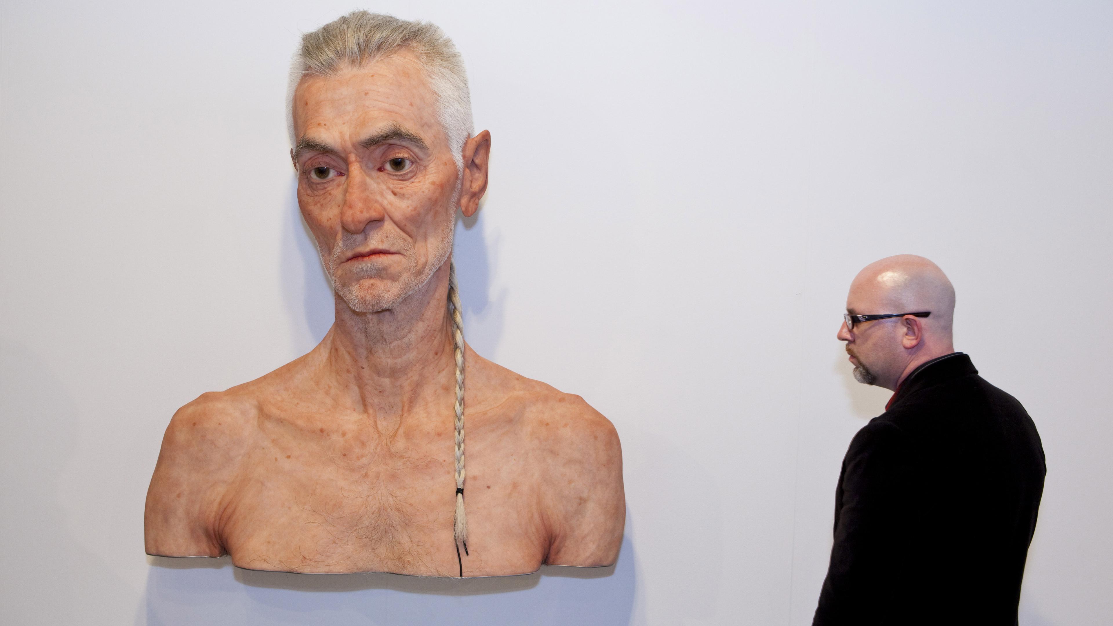 A man observing a large, lifelike sculpture of an elderly man with a braid on a white gallery wall.