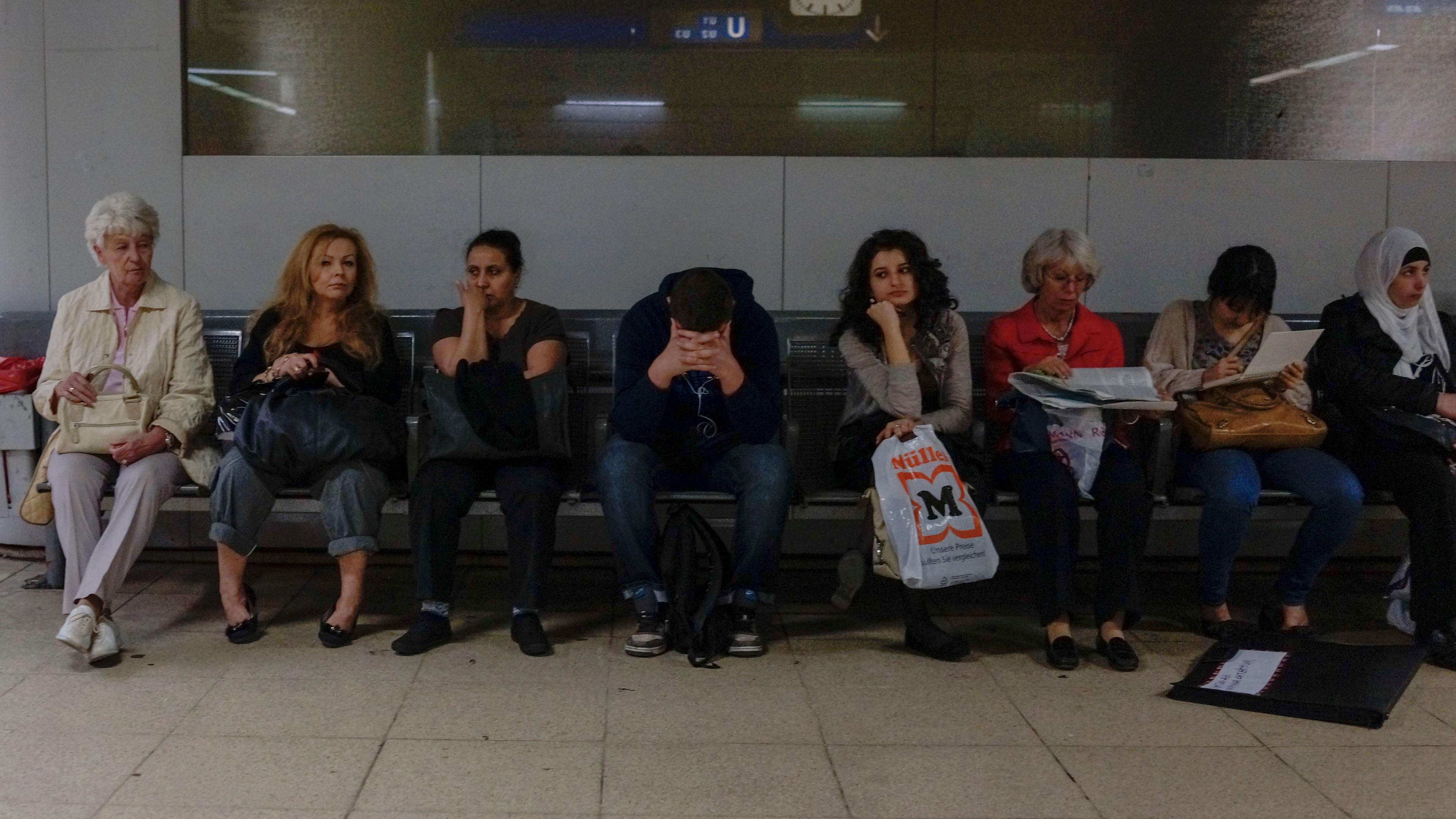 Photo of seven people sitting on a bench in a train station waiting area, with a clock on the wall behind them.