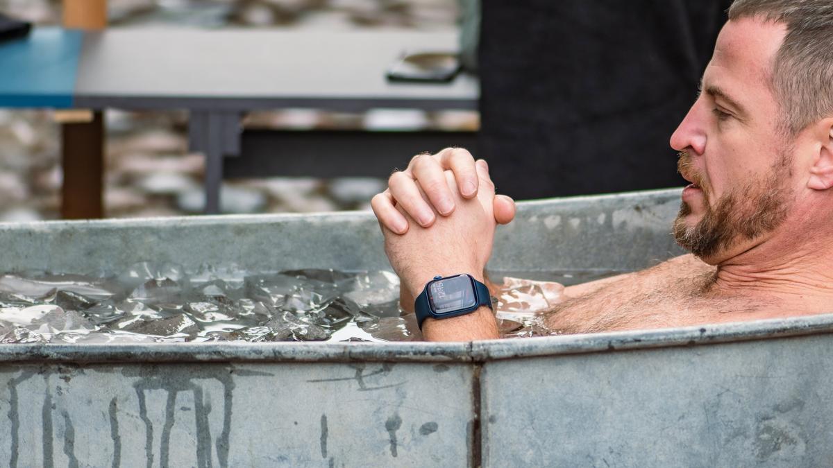 A man in an outdoor ice bath, clasping his hands and wearing a smartwatch.