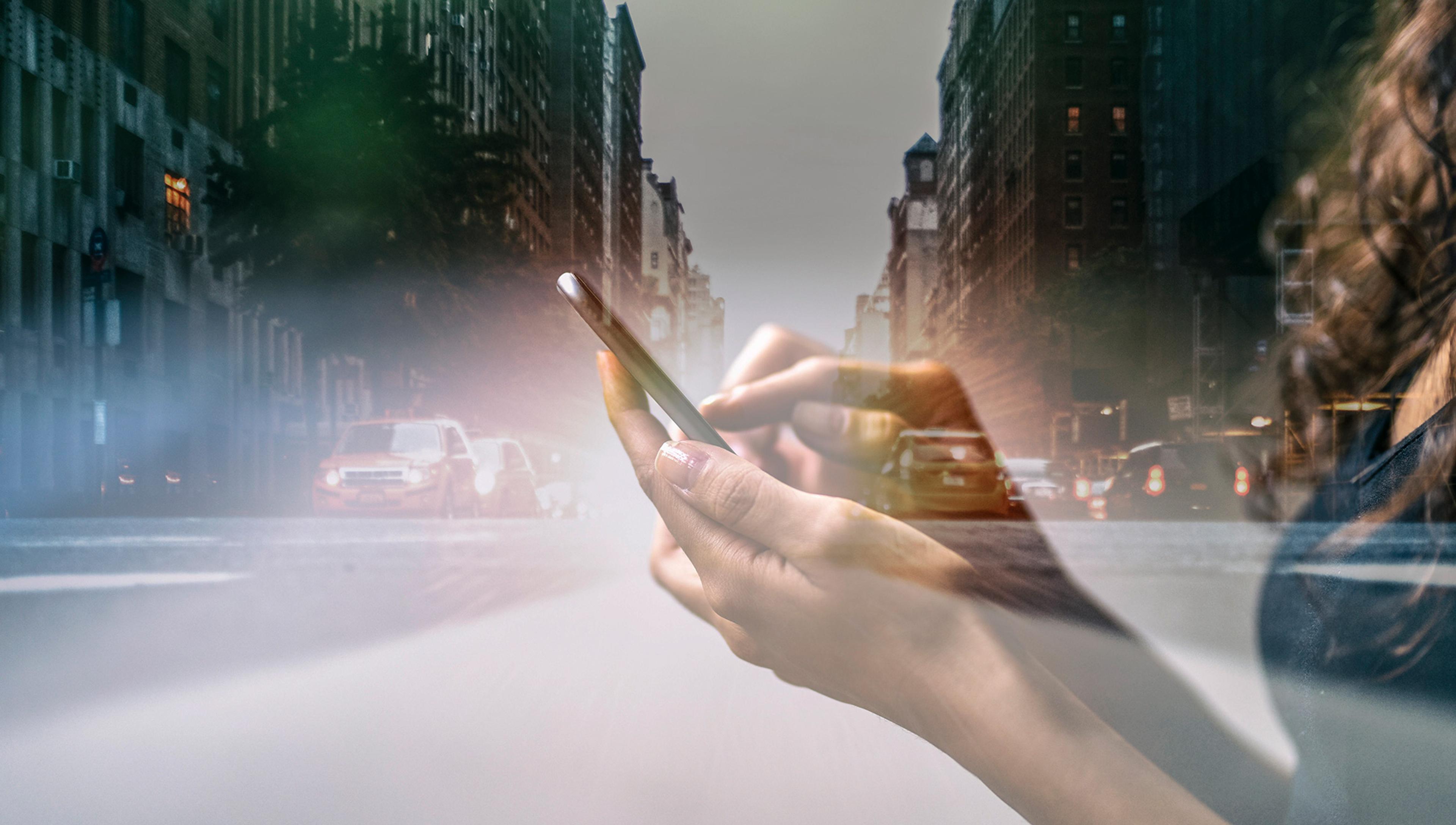 A double exposure image of a person using a smartphone overlaid with a bustling city street scene with cars and tall buildings.