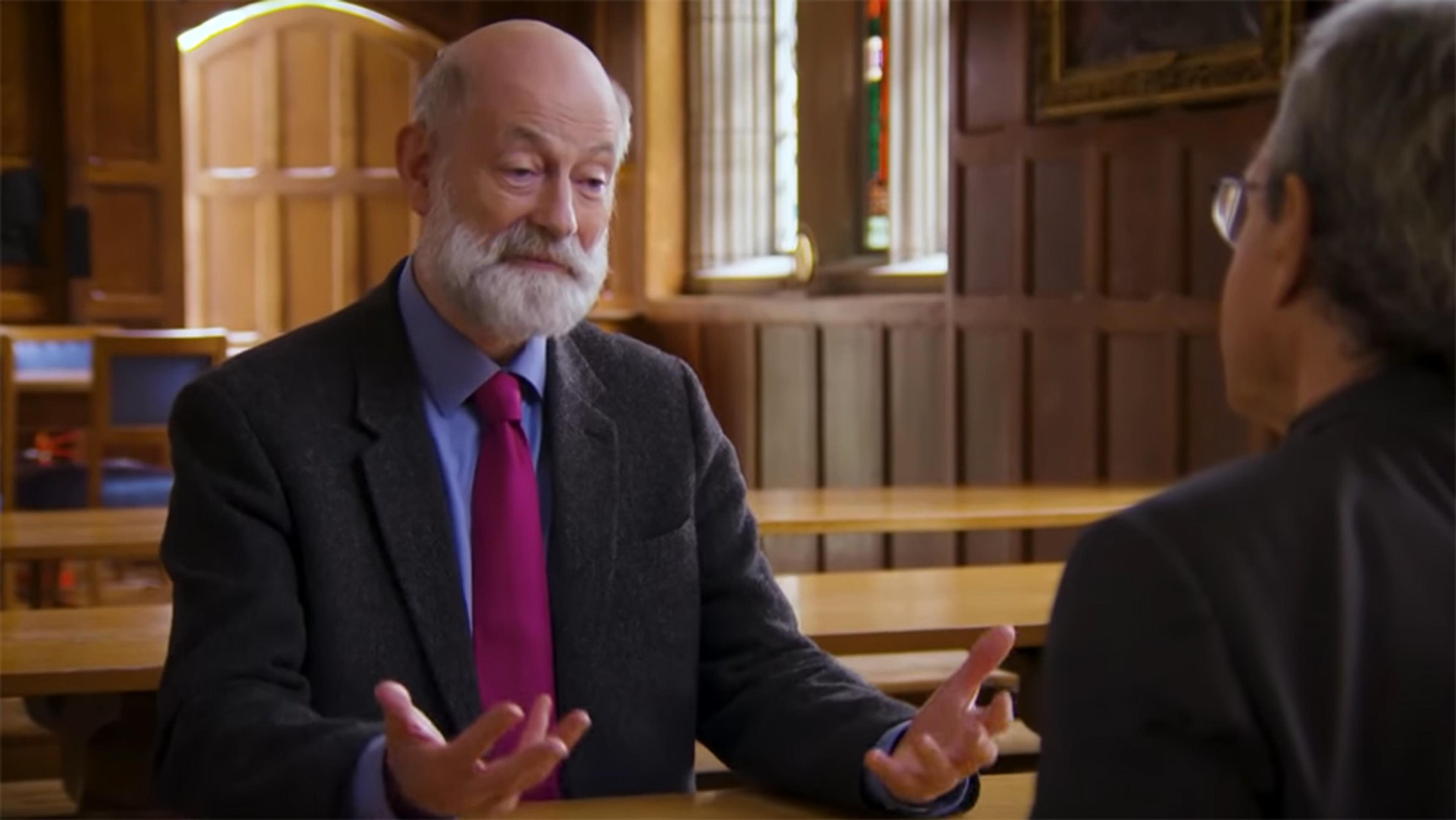 Two men talking in a wooden-panelled room with stained glass windows. One bald man with a grey beard gestures with his hands while speaking.