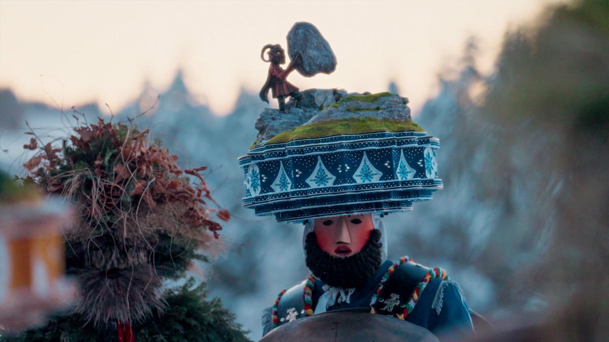 Photo of a person in a decorative costume with a mask and an elaborate headpiece featuring a figure lifting a rock.