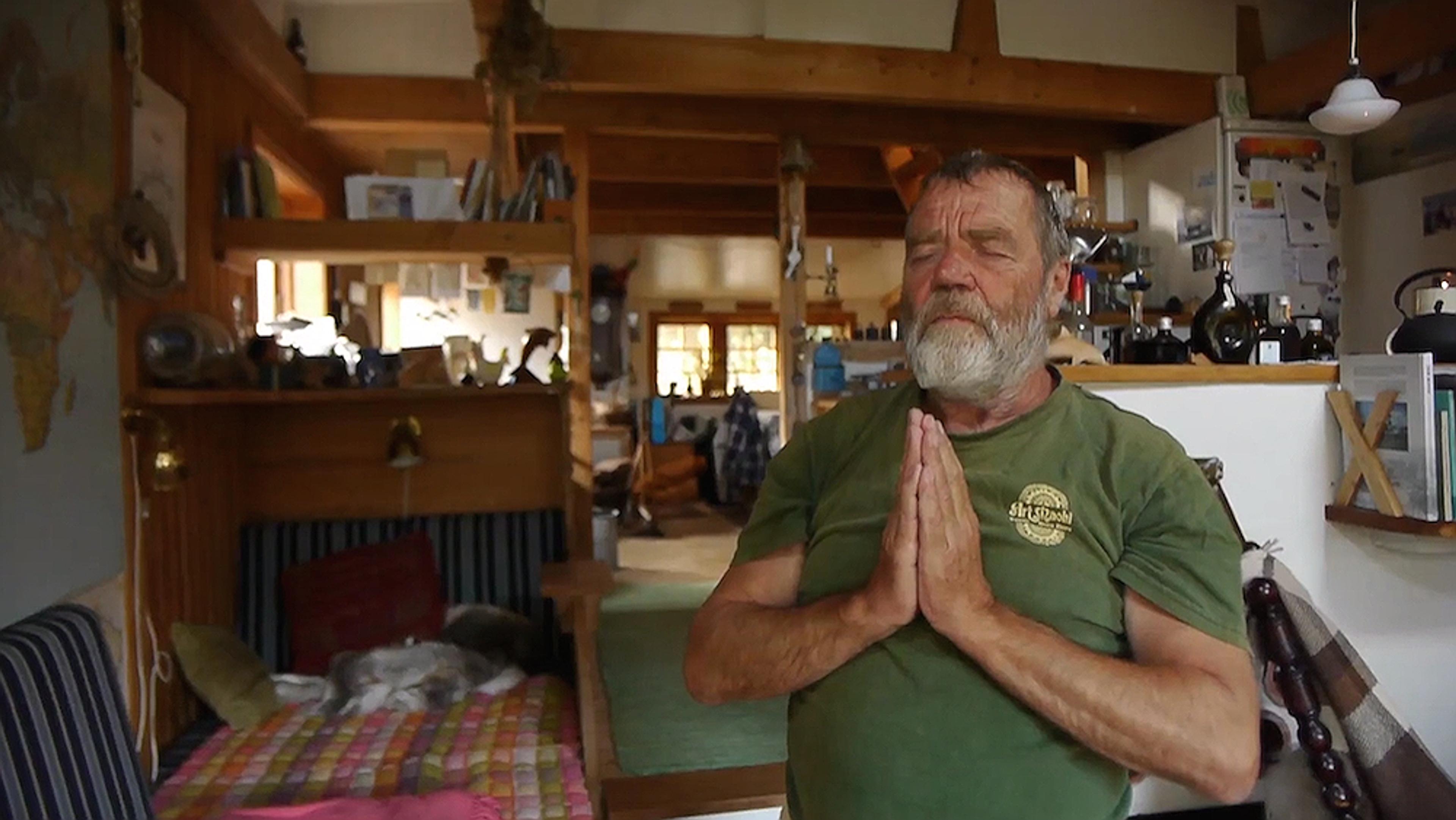 A man with closed eyes and hands in prayer pose inside a cosy, cluttered wooden house with various decorations and furniture.