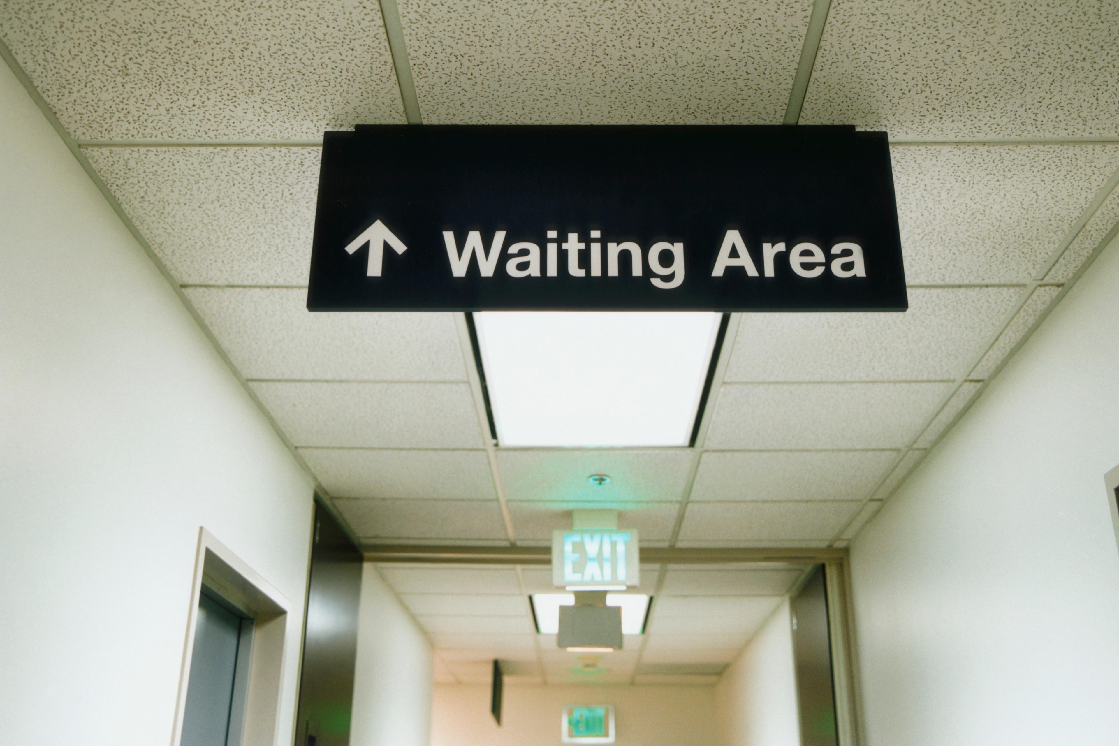 Photo of a hospital corridor with a ceiling sign pointing to the waiting area. An exit sign is visible in the background.