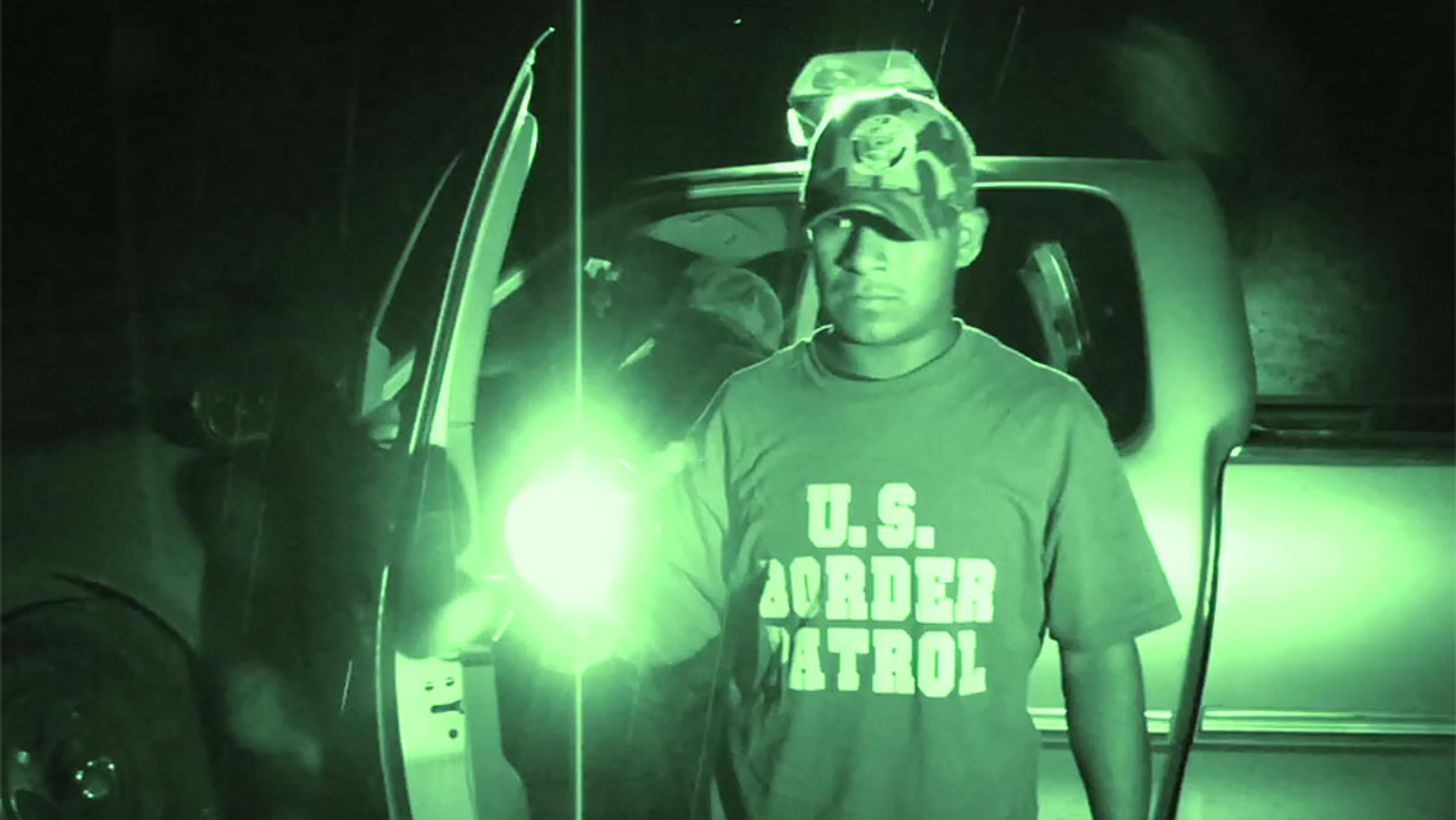 Night vision photo of a person wearing a U.S. Border Patrol shirt standing next to an open car door holding a light.