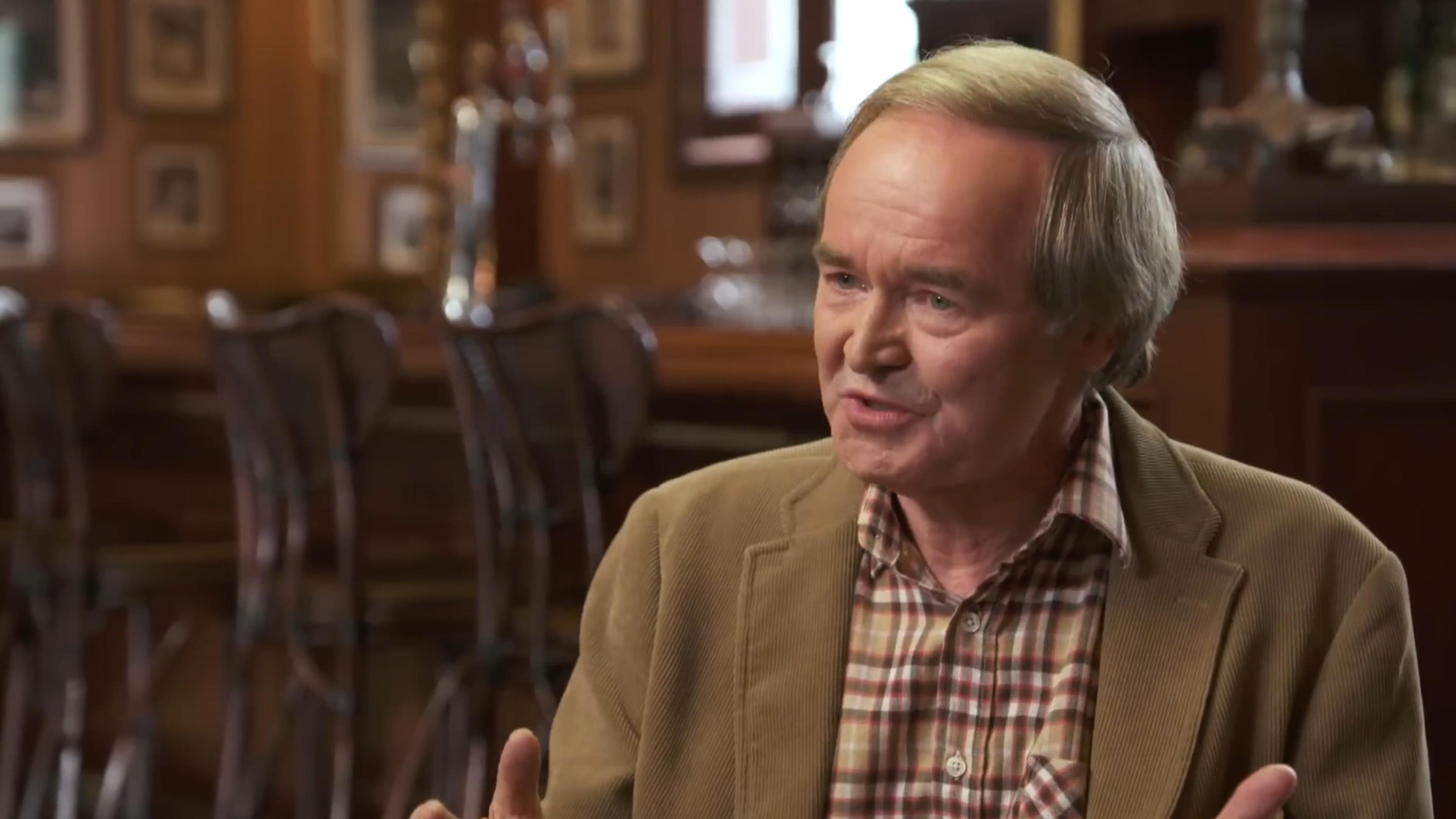 Photo of an older man in a checked shirt and brown jacket speaking, bar with framed photos in the background.
