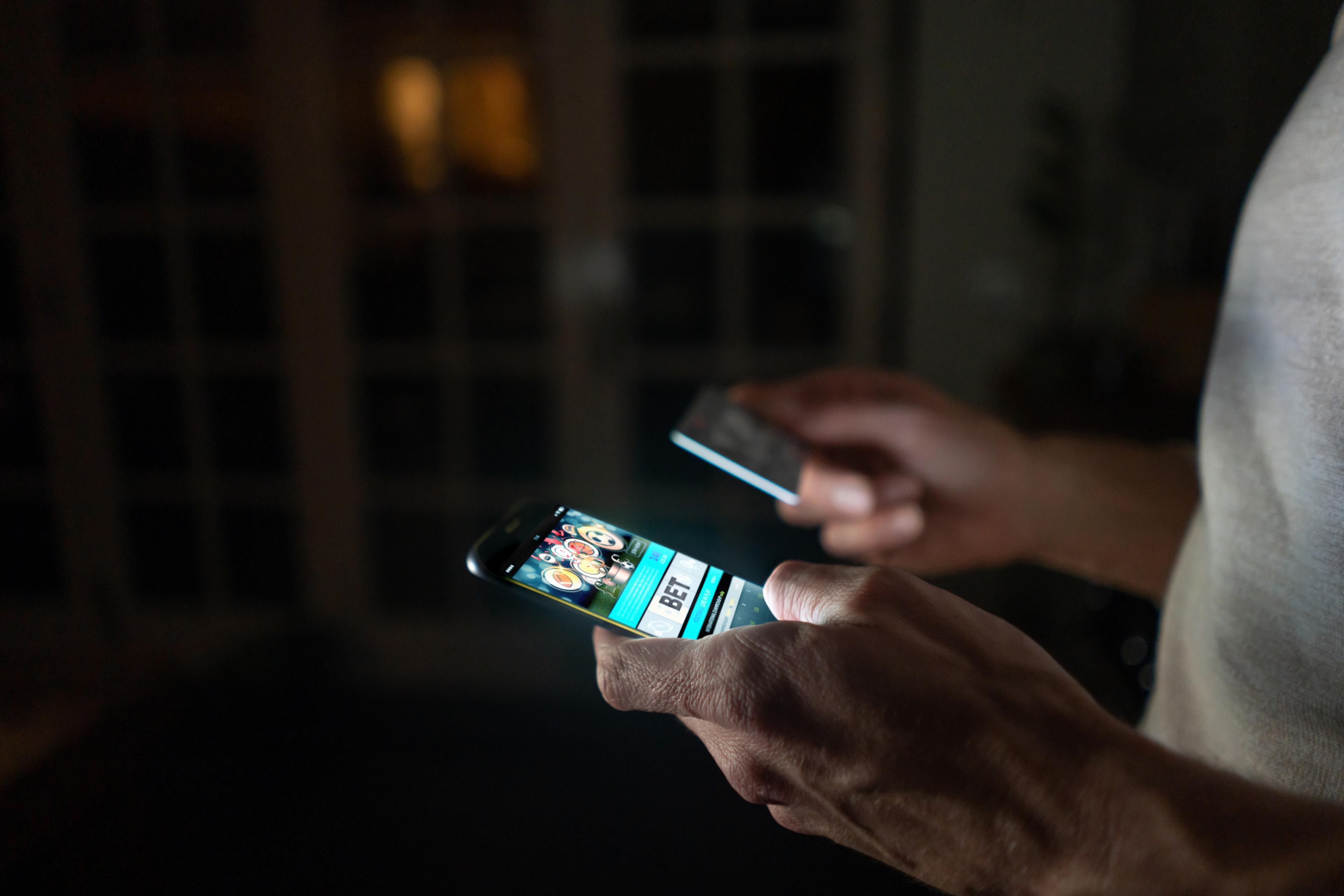 Photo of a person in a dim room holding a smartphone and a credit card, phone screen showing a betting app.
