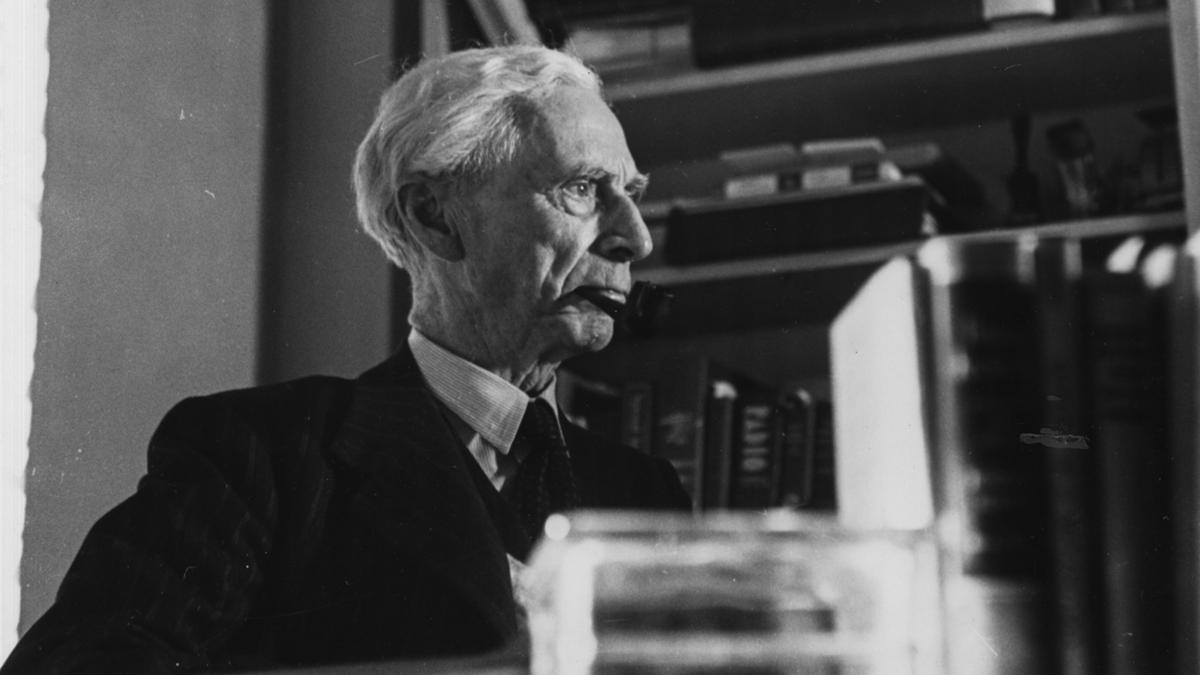 Black and white photo of an elderly man with a pipe, sitting in front of bookshelves filled with books and stacks.