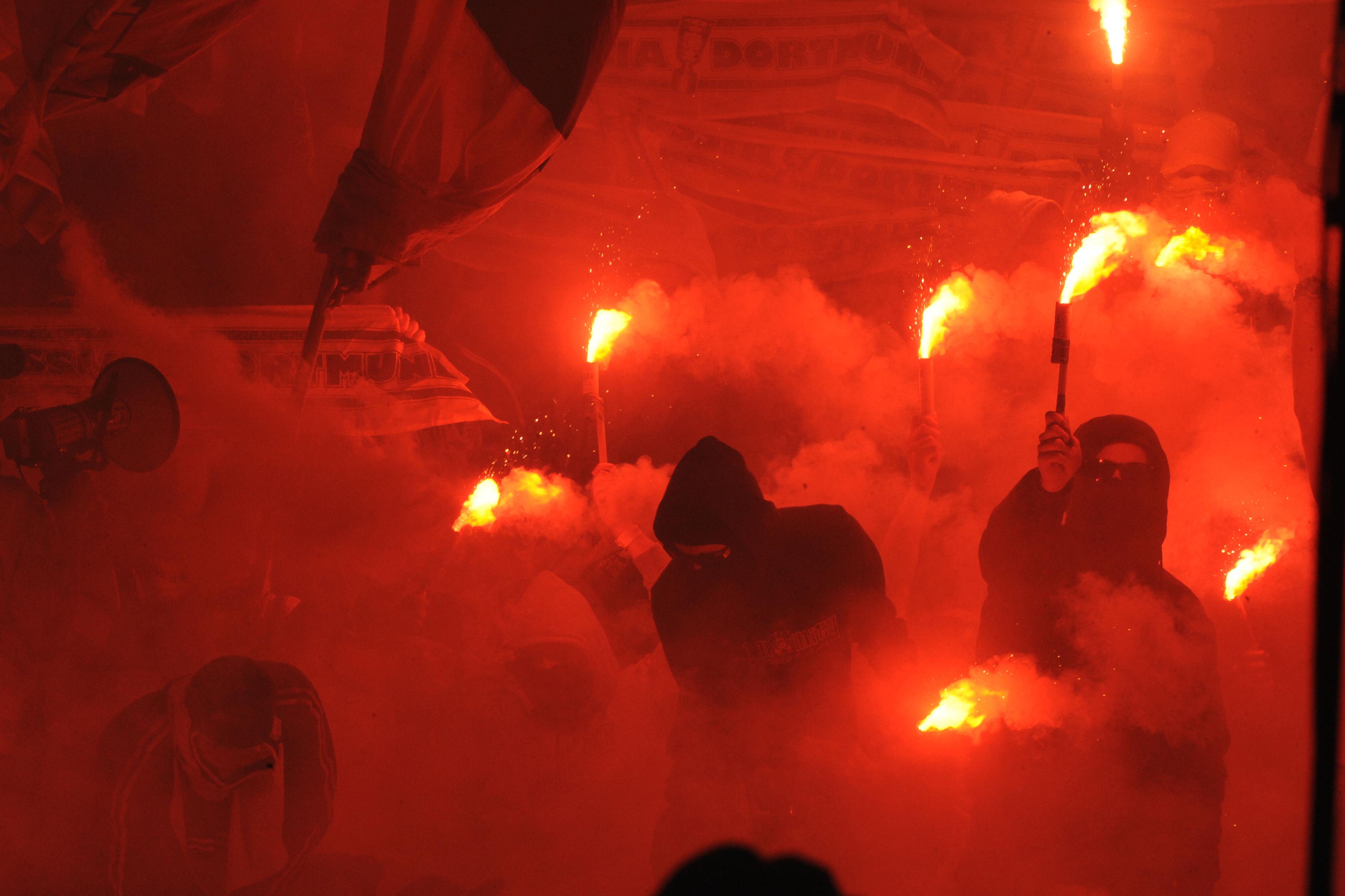 Photo of people in hoodies holding flares, surrounded by smoke and red light, conveying intense atmosphere.