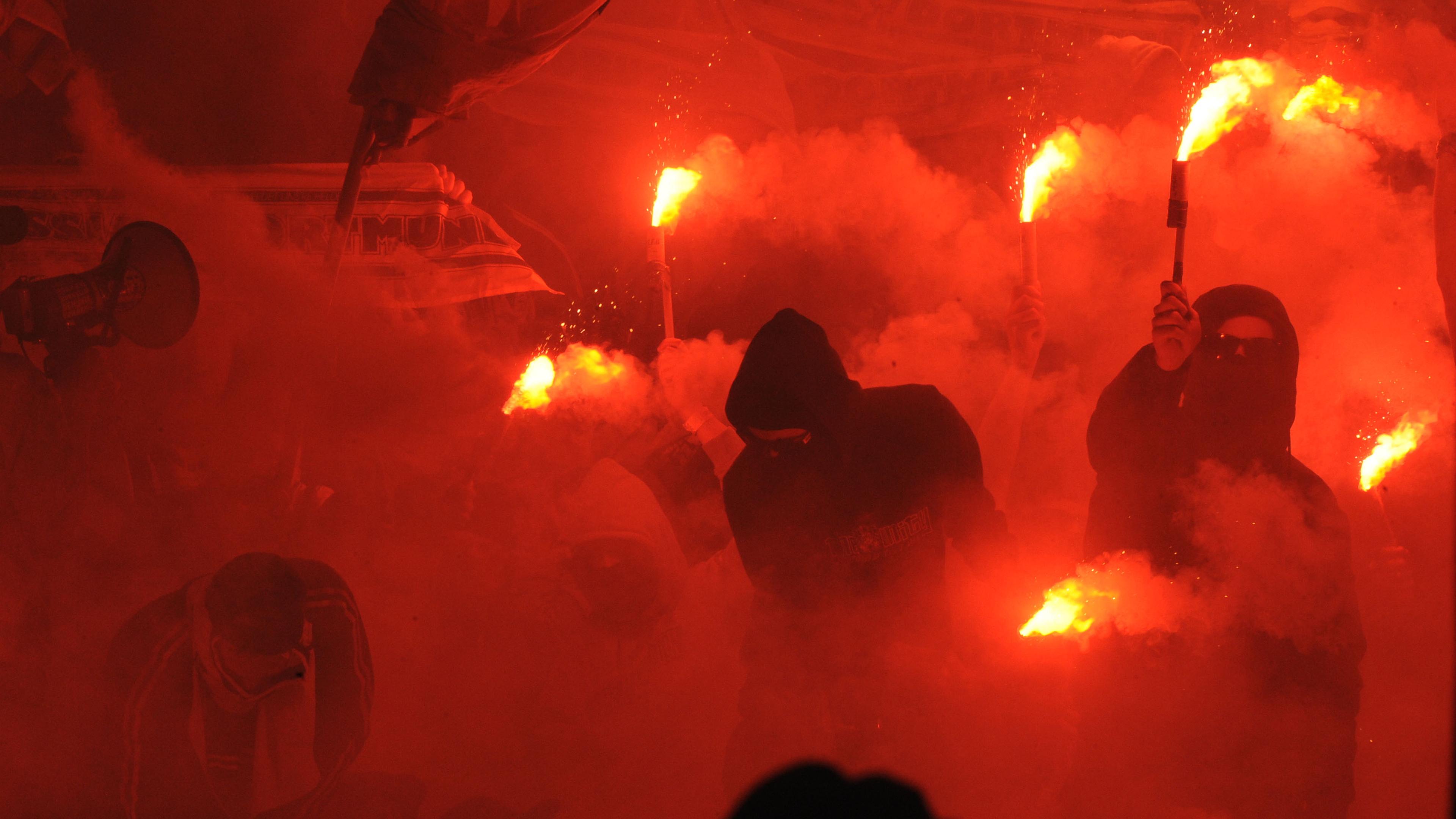 Photo of people in hoodies holding flares, surrounded by smoke and red light, conveying intense atmosphere.