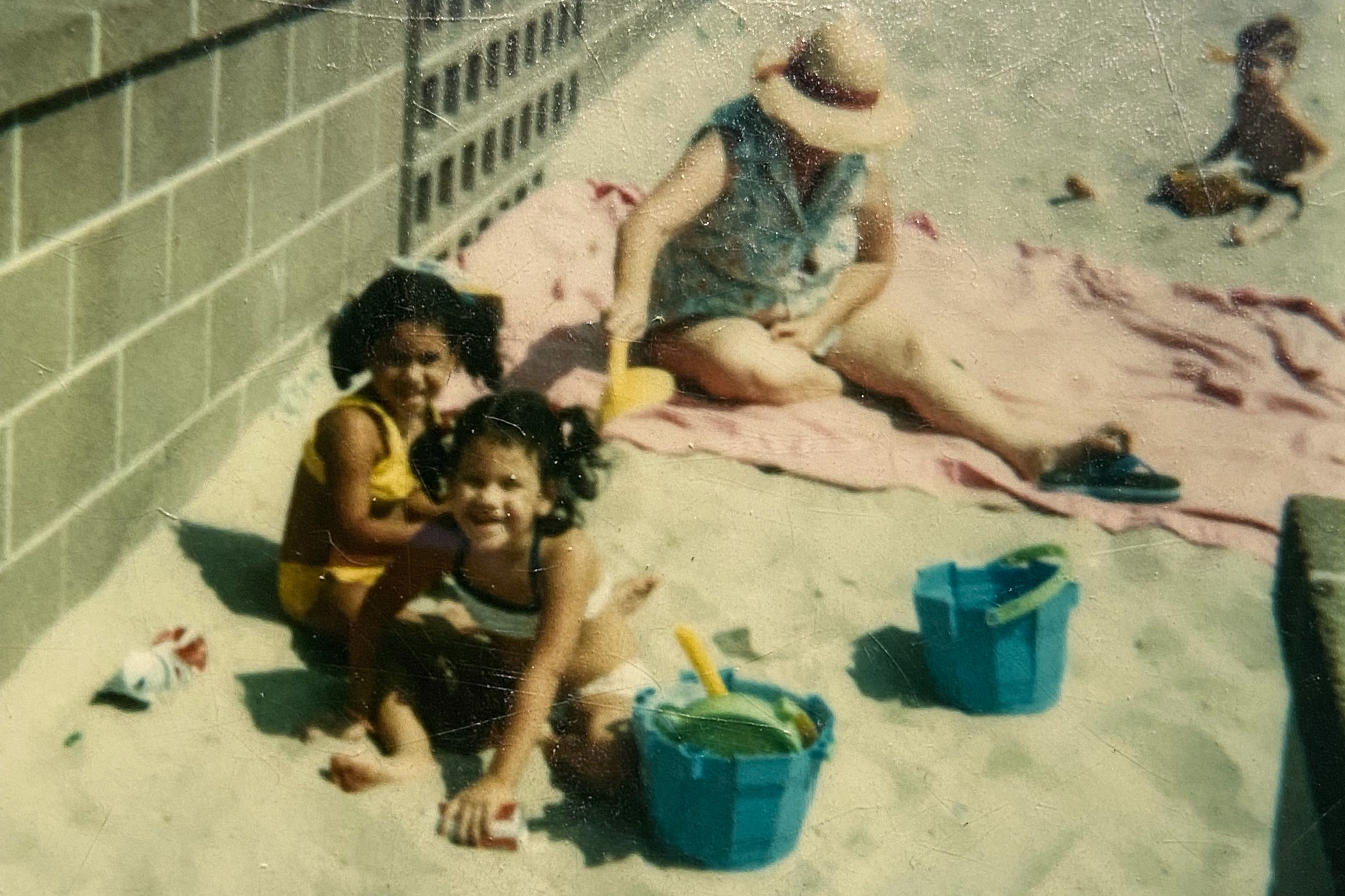 Children playing with buckets in sand by a brick wall, an adult sits on a towel wearing a hat nearby.
