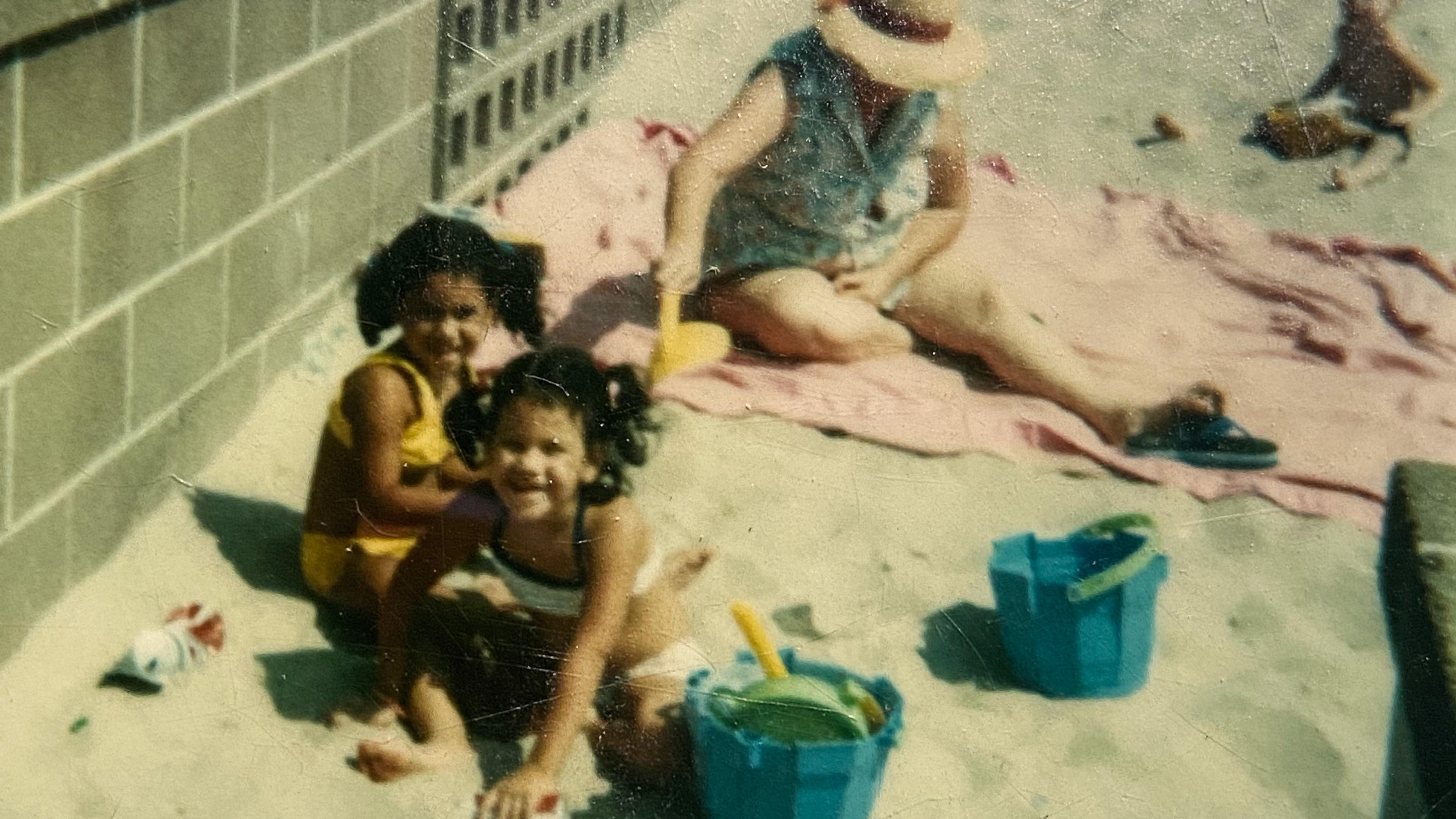 Children playing with buckets in sand by a brick wall, an adult sits on a towel wearing a hat nearby.