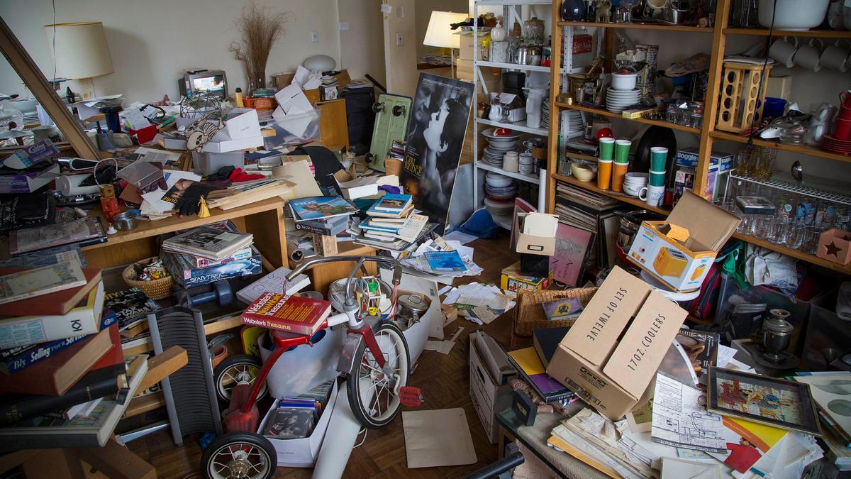 Photo of a cluttered room filled with scattered books, papers, boxes and household items on shelves and floor.