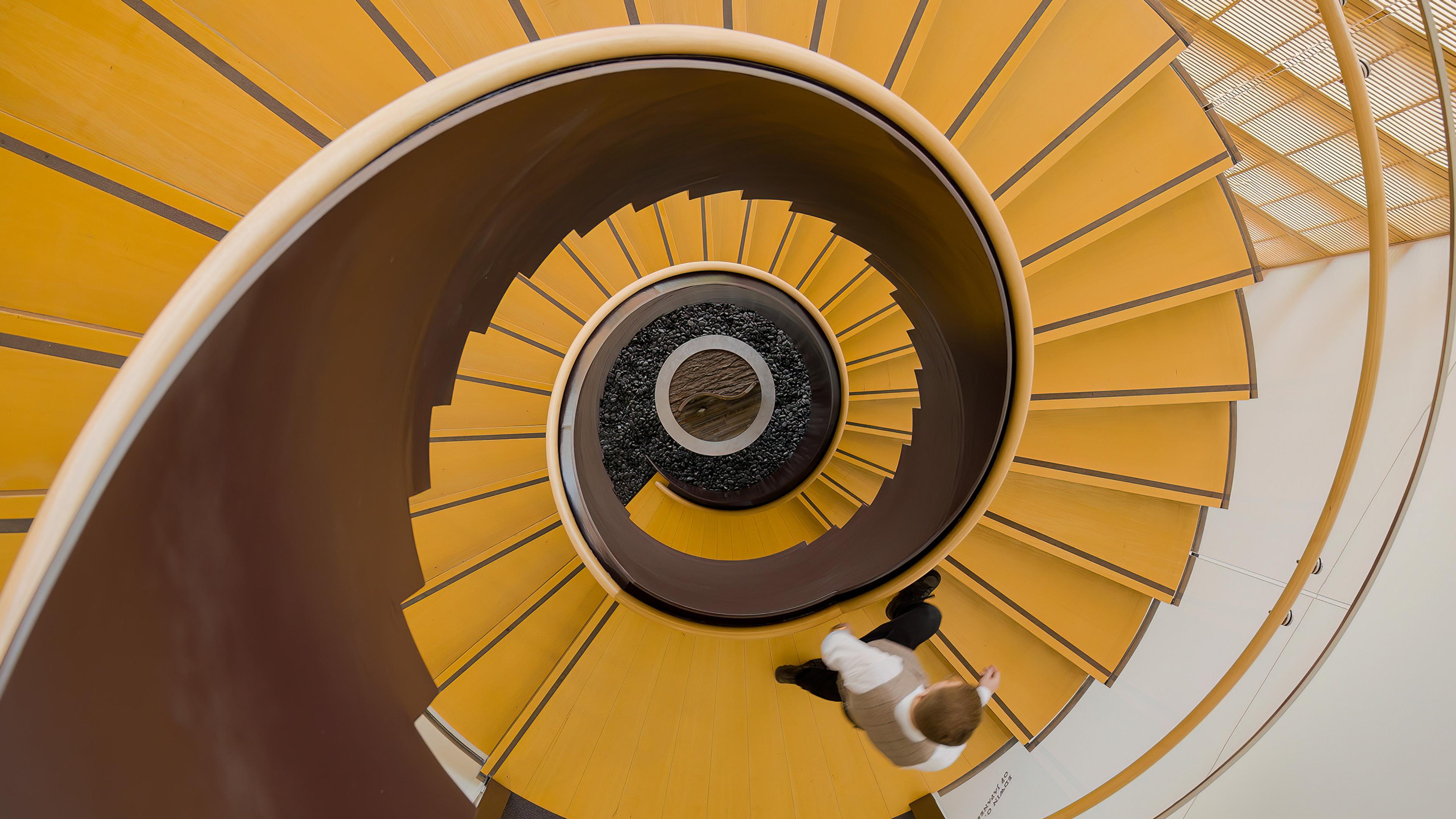 Overhead view of a person walking down a spiral staircase with yellow steps. The staircase creates a swirling visual effect.