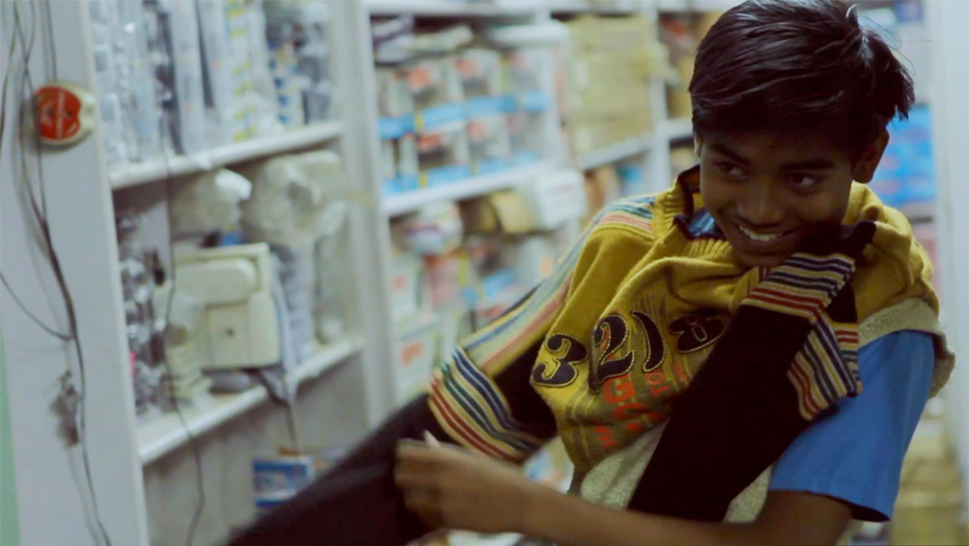 A boy smiling while putting on a striped jumper in a shop with shelves filled with various goods in the background.