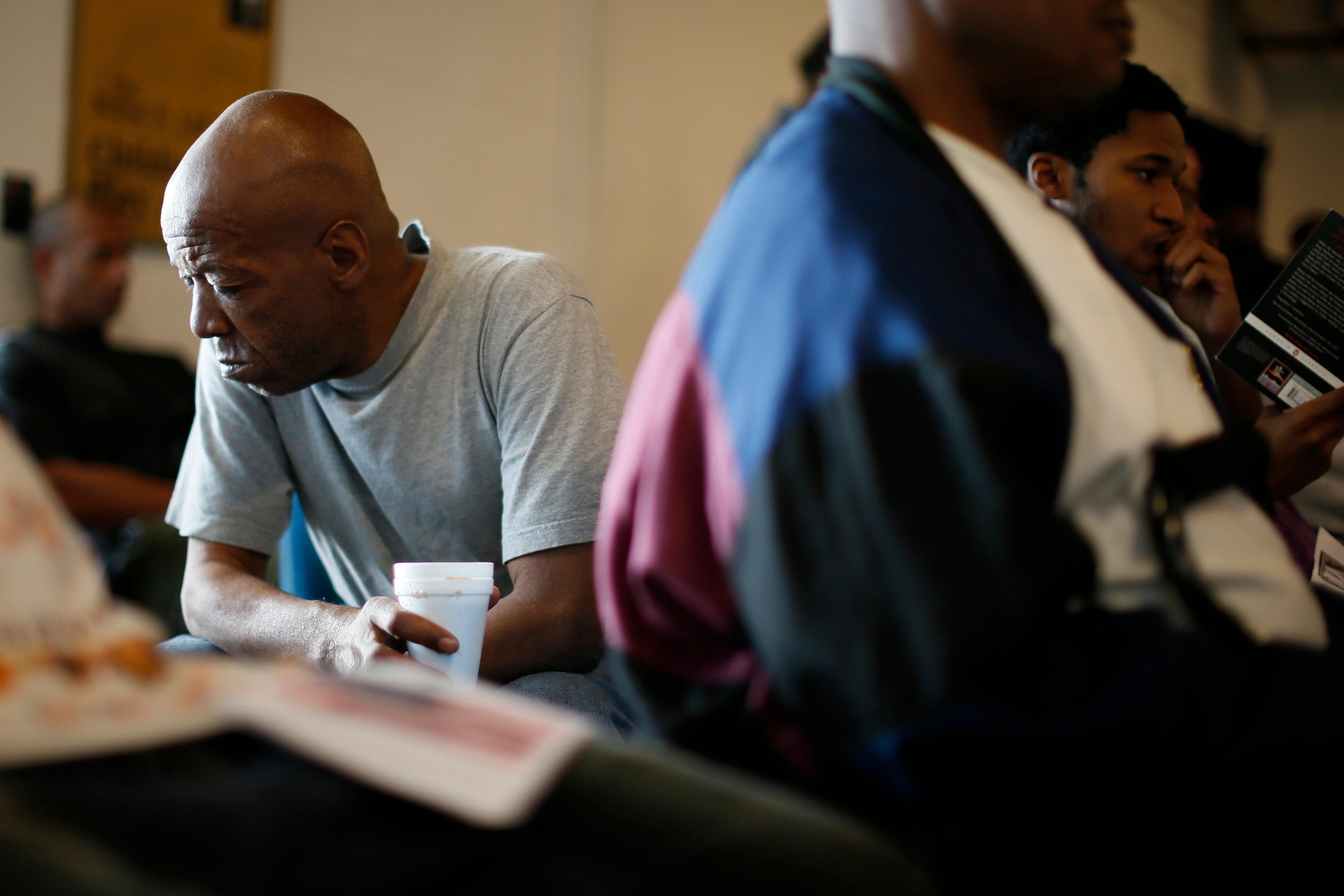 Photo of a man in a grey shirt holding a cup, sitting among others in a room, with a pensive expression on his face.
