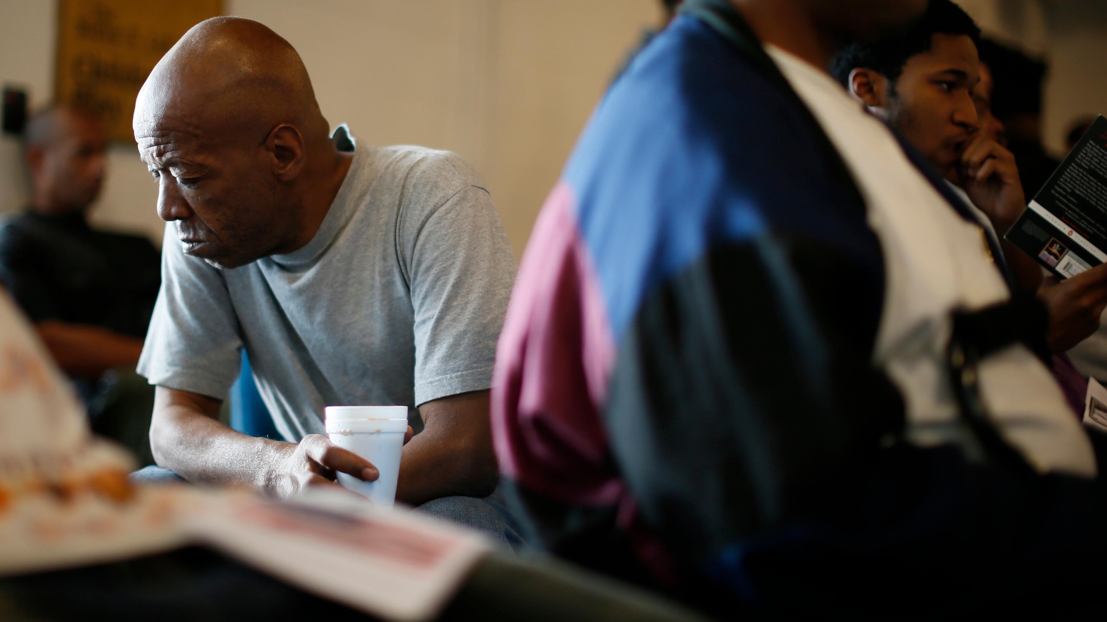 Photo of a man in a grey shirt holding a cup, sitting among others in a room, with a pensive expression on his face.