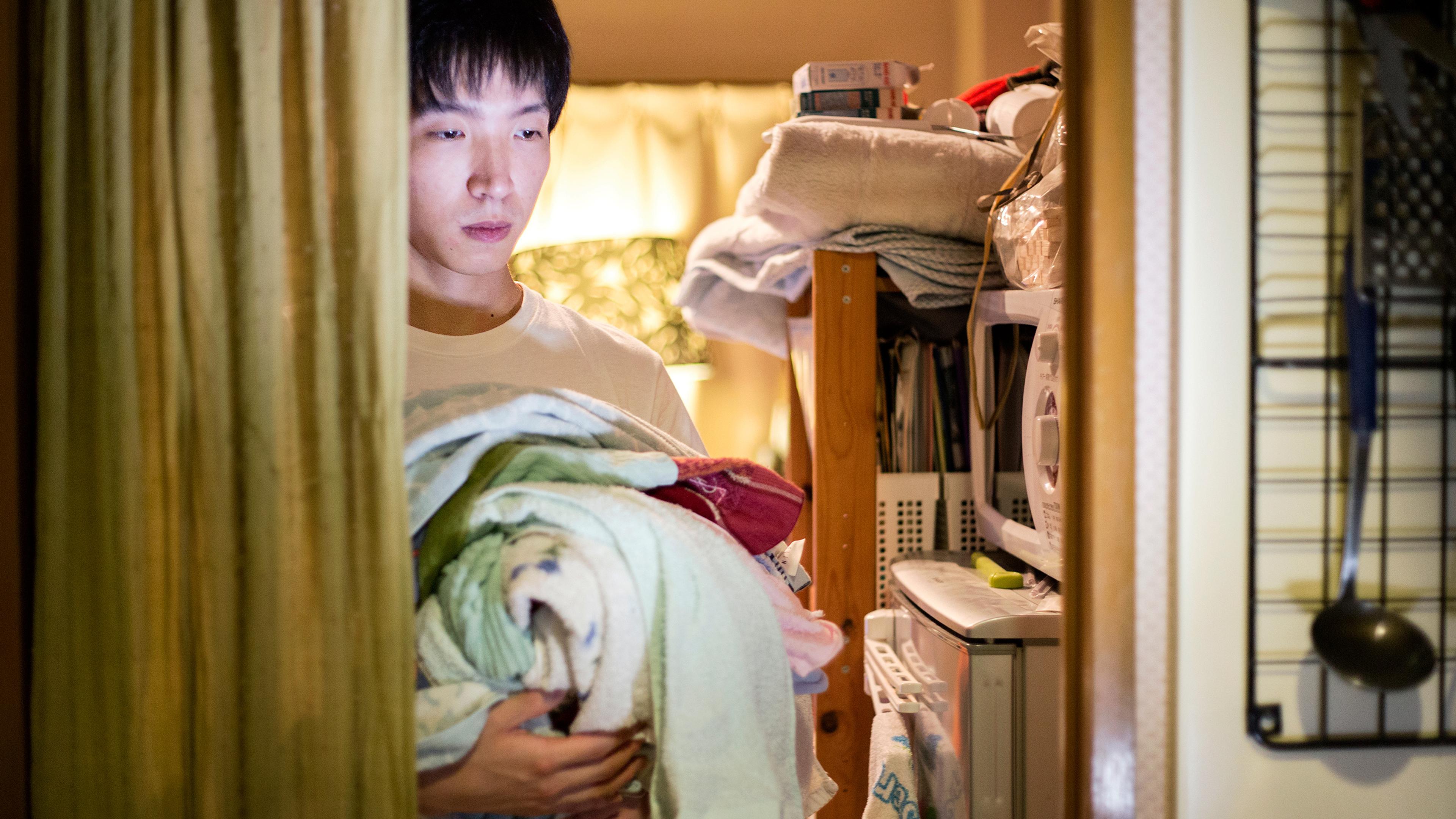 Photo of a man holding laundry in a dimly lit room with shelves, a microwave and a curtain on the left.