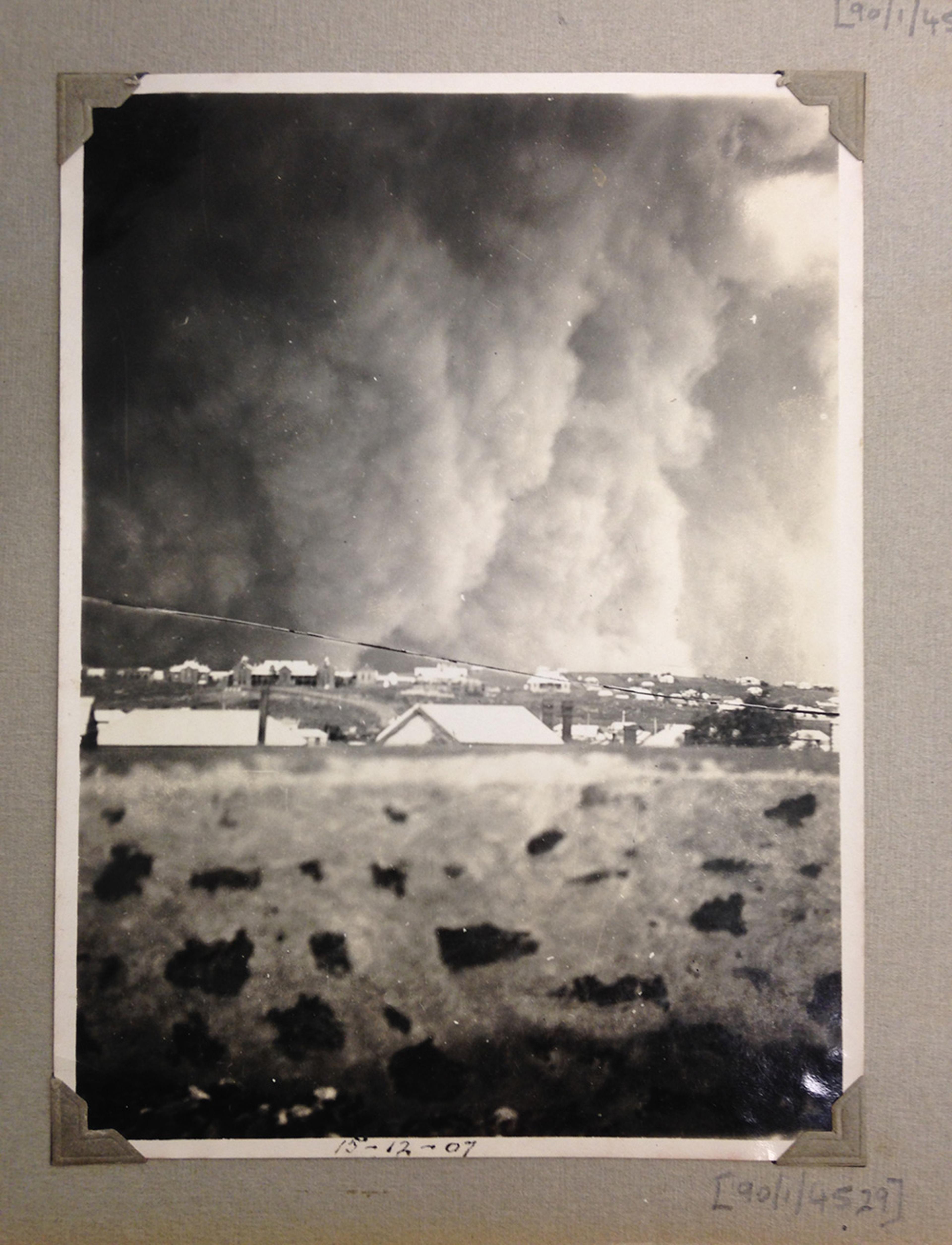 Black and white photo of a townscape with large plumes of smoke or dust rising in the sky, dated 13-12-07 in the lower centre.