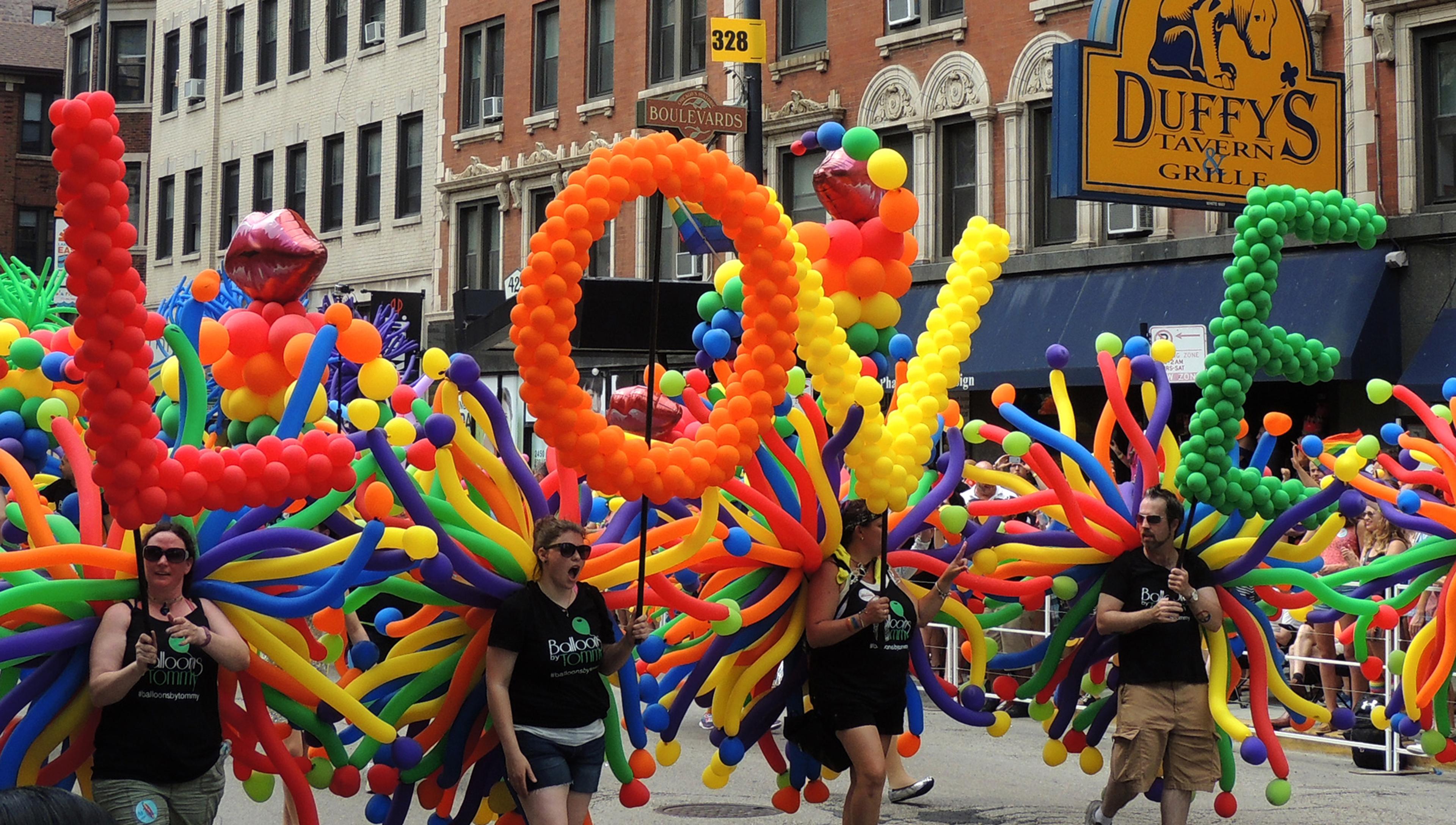 Participants in a parade carry large balloon structures spelling “LOVE” in bright colours, with spectators in the background.