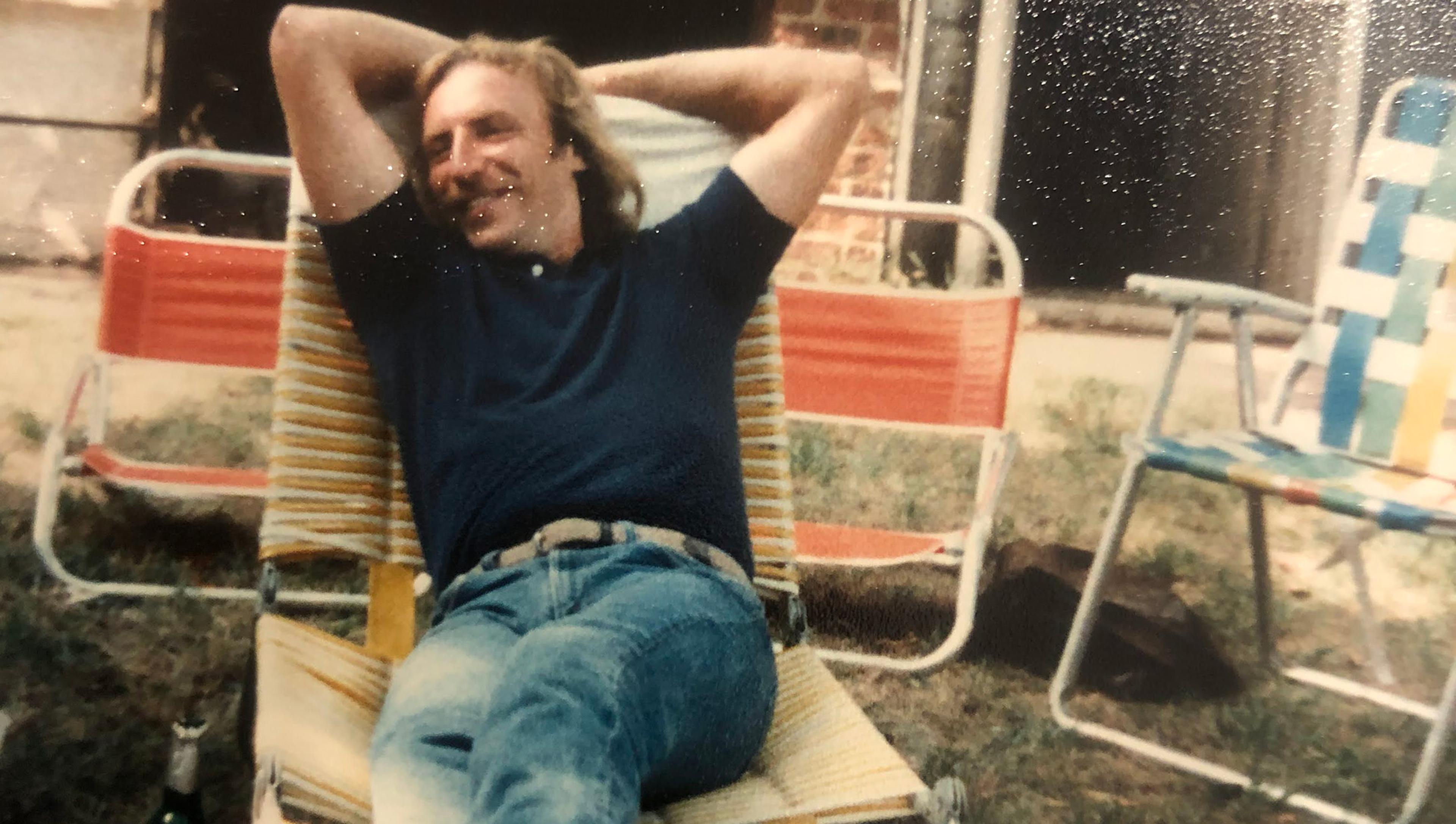 Man relaxing on a striped lounge chair with hands behind his head, outside on a lawn, flanked by other coloured chairs and an opened beer bottle nearby.