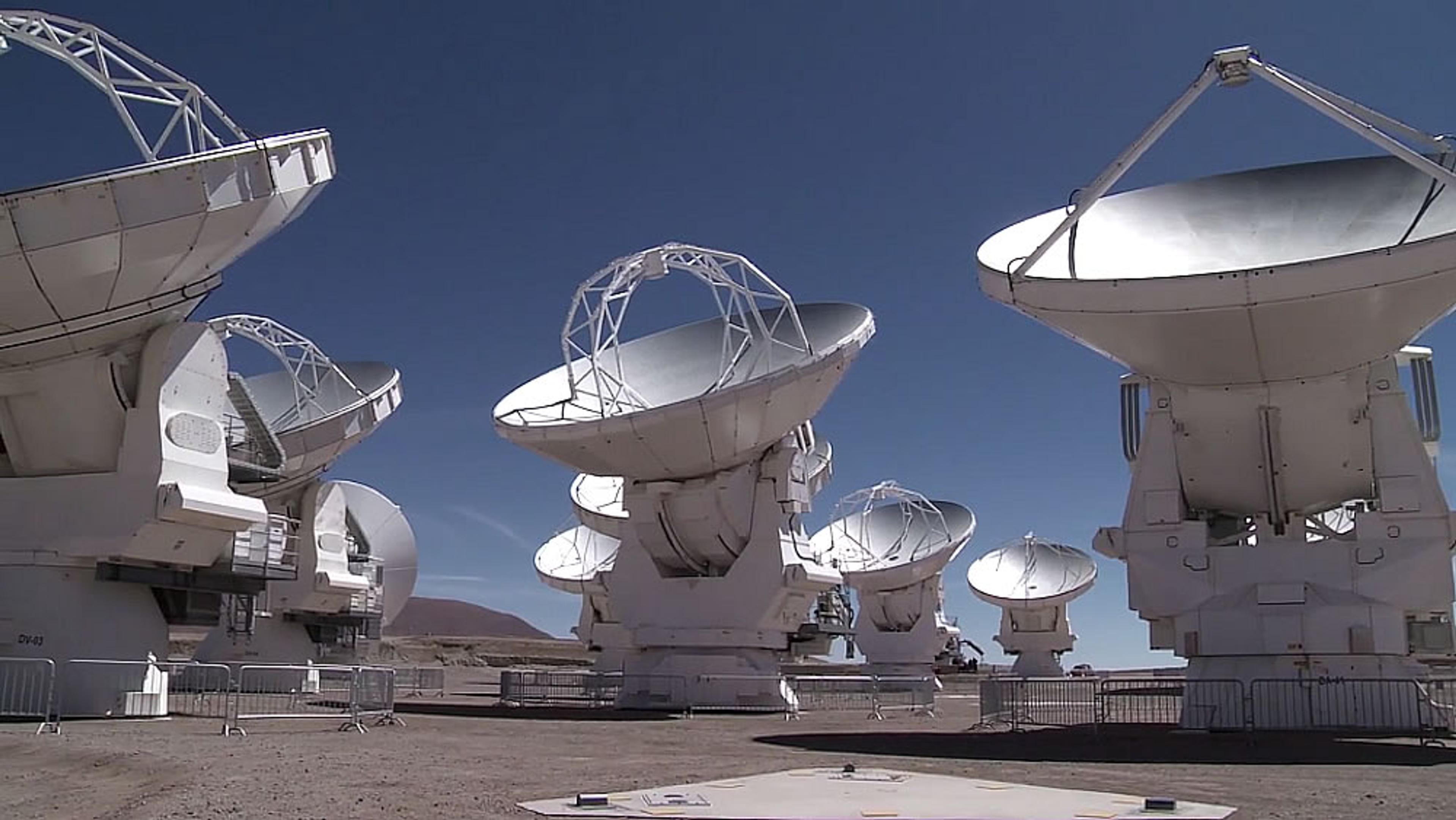 Multiple large radio telescopes with white dishes under a clear blue sky in a desolate, fenced area.