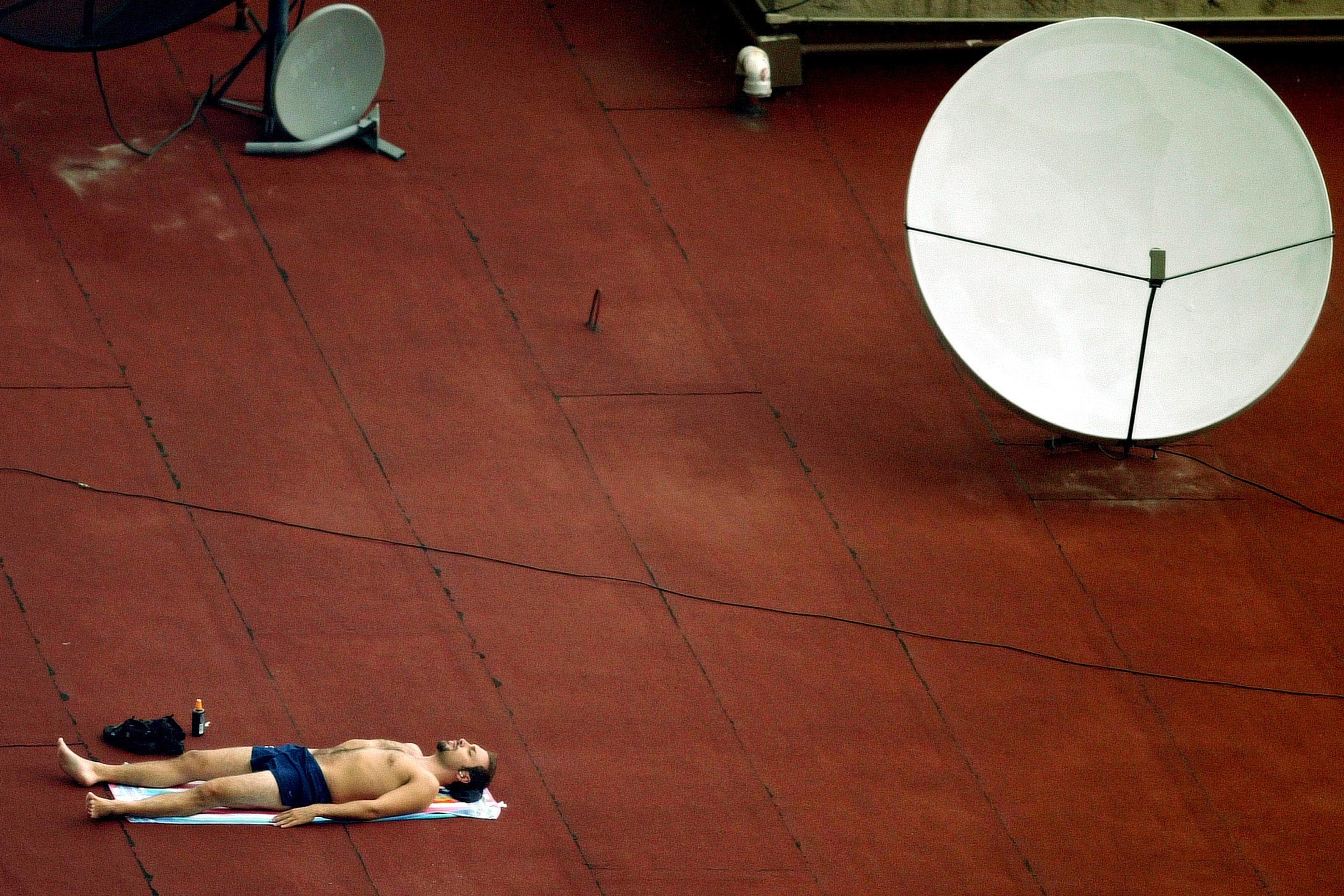Photo of a man sunbathing on a rooftop next to a large satellite dish.