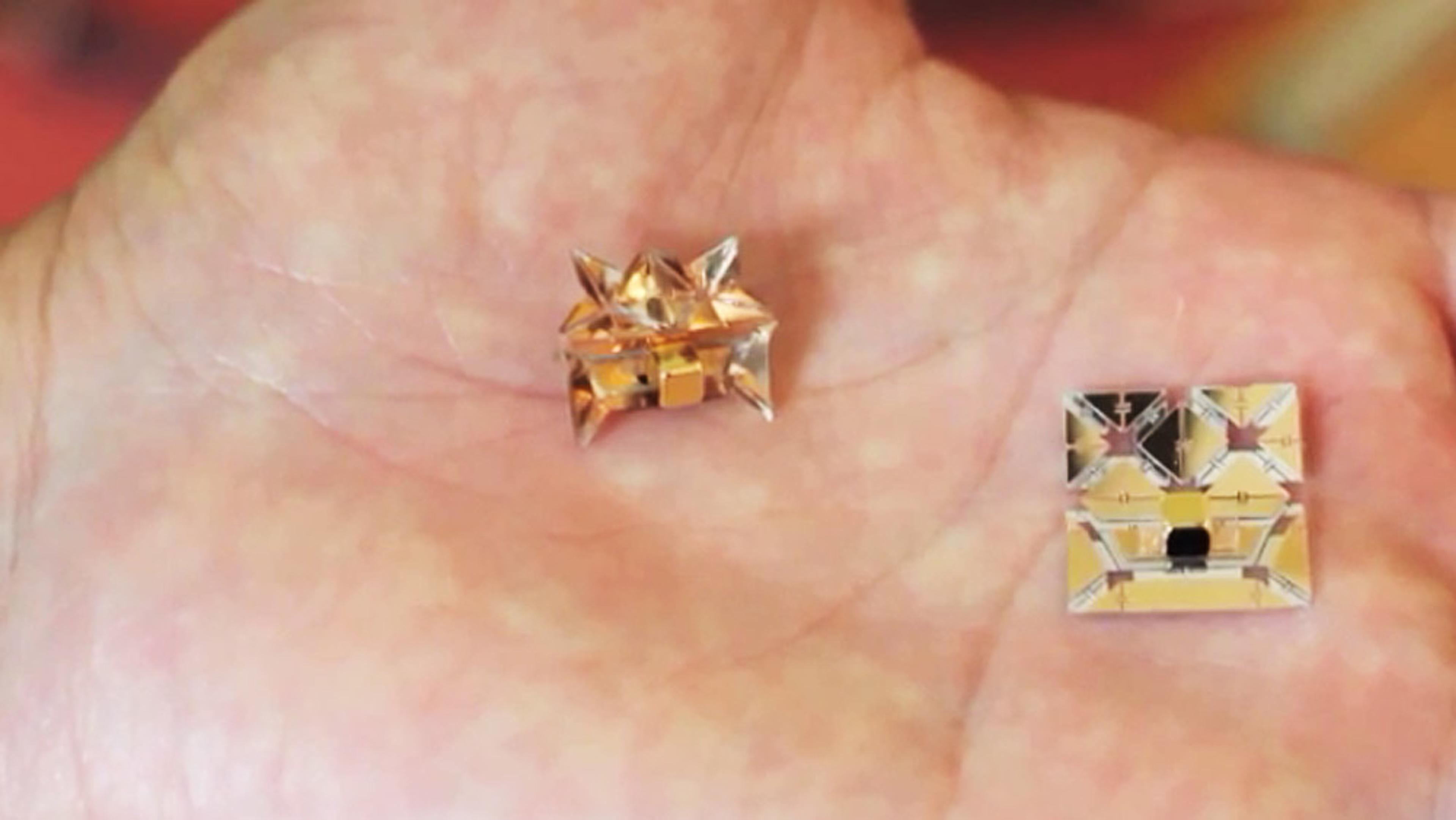 Close-up of a hand holding a tiny gold-coloured origami robot and its flat, unfolded counterpart.
