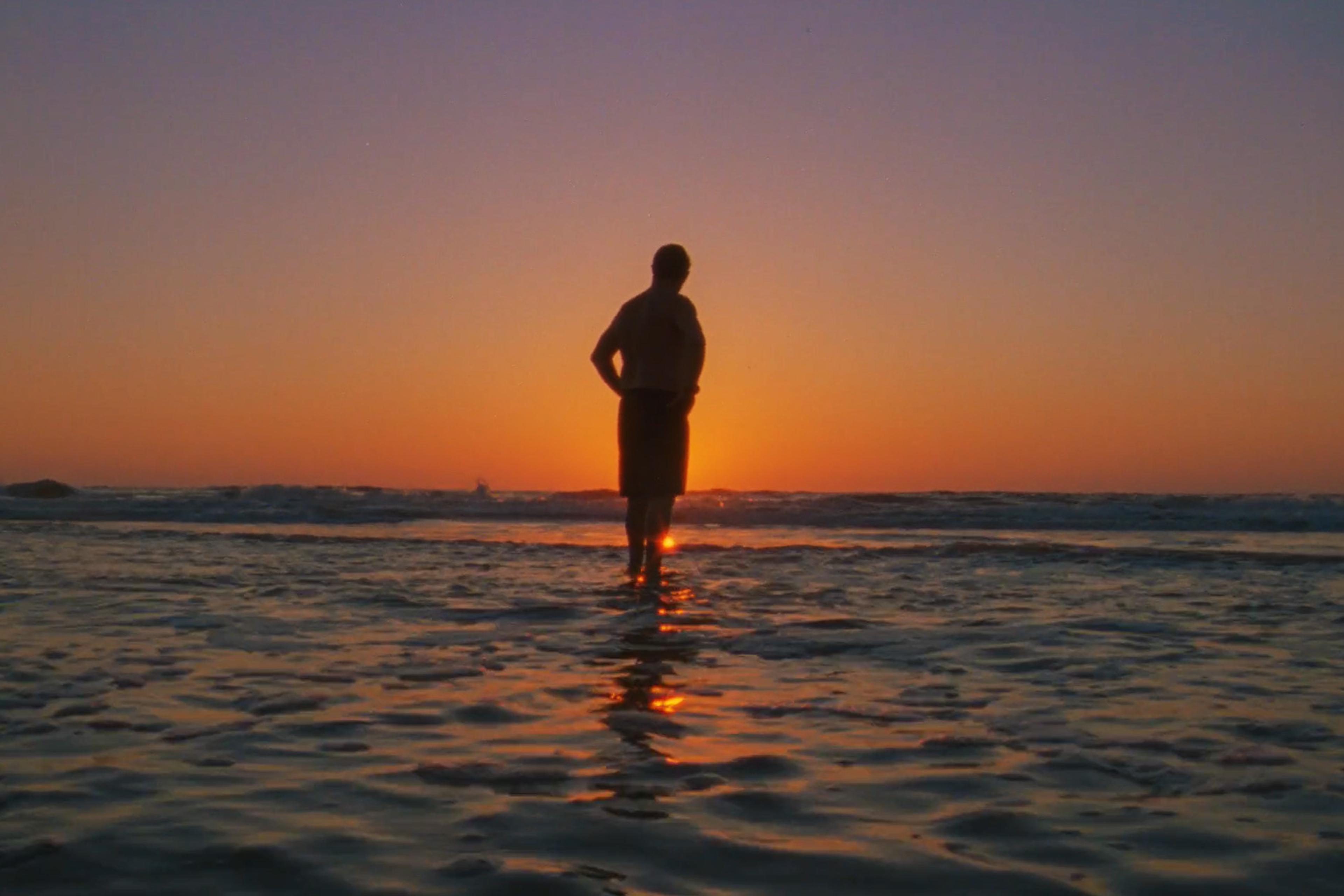 A person stands on the shore, watching the sunset over the ocean, with the sky in vibrant shades of orange and purple.