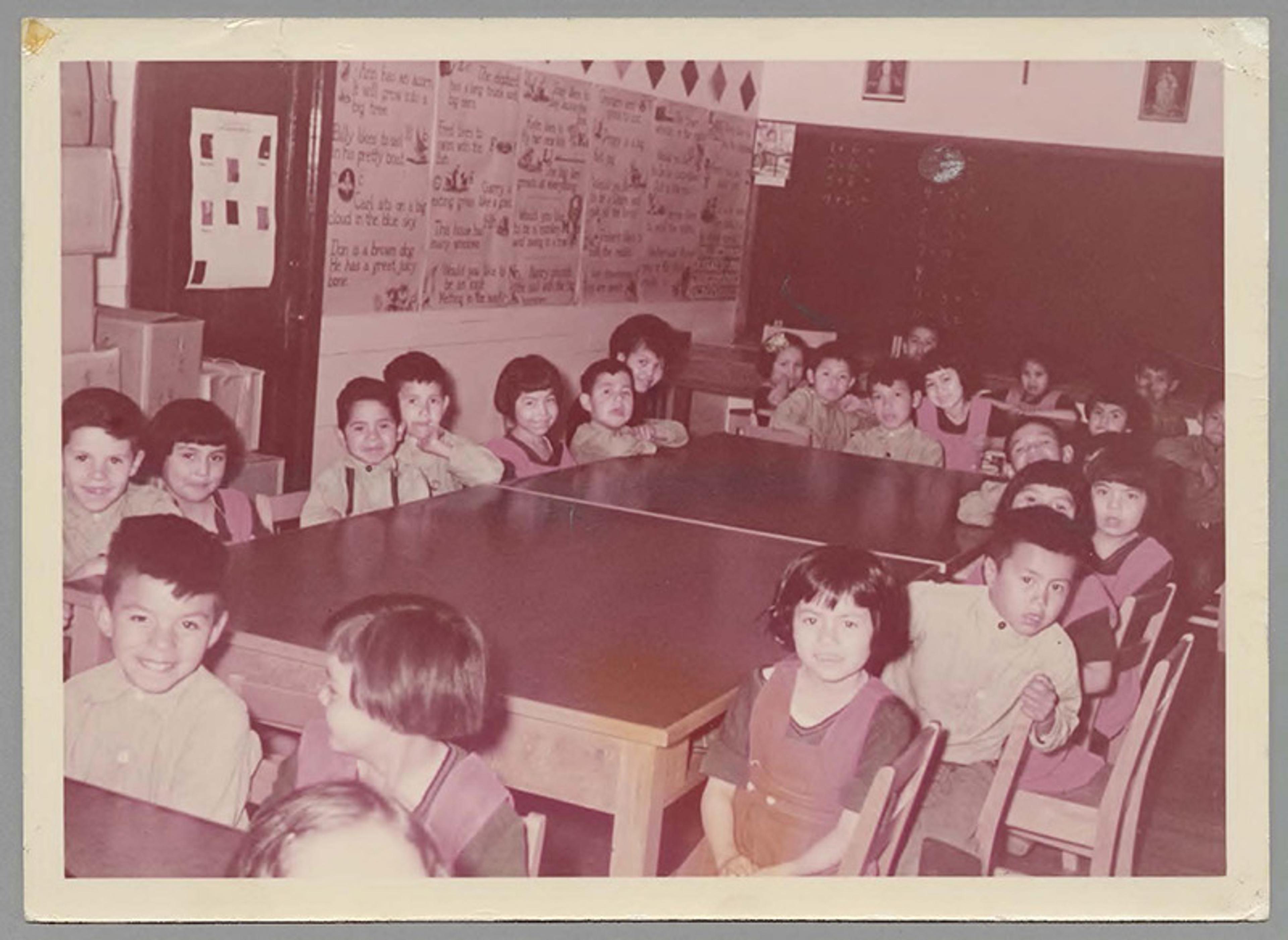 Vintage photo of young children seated around tables in a classroom, with educational posters on the wall in the background.