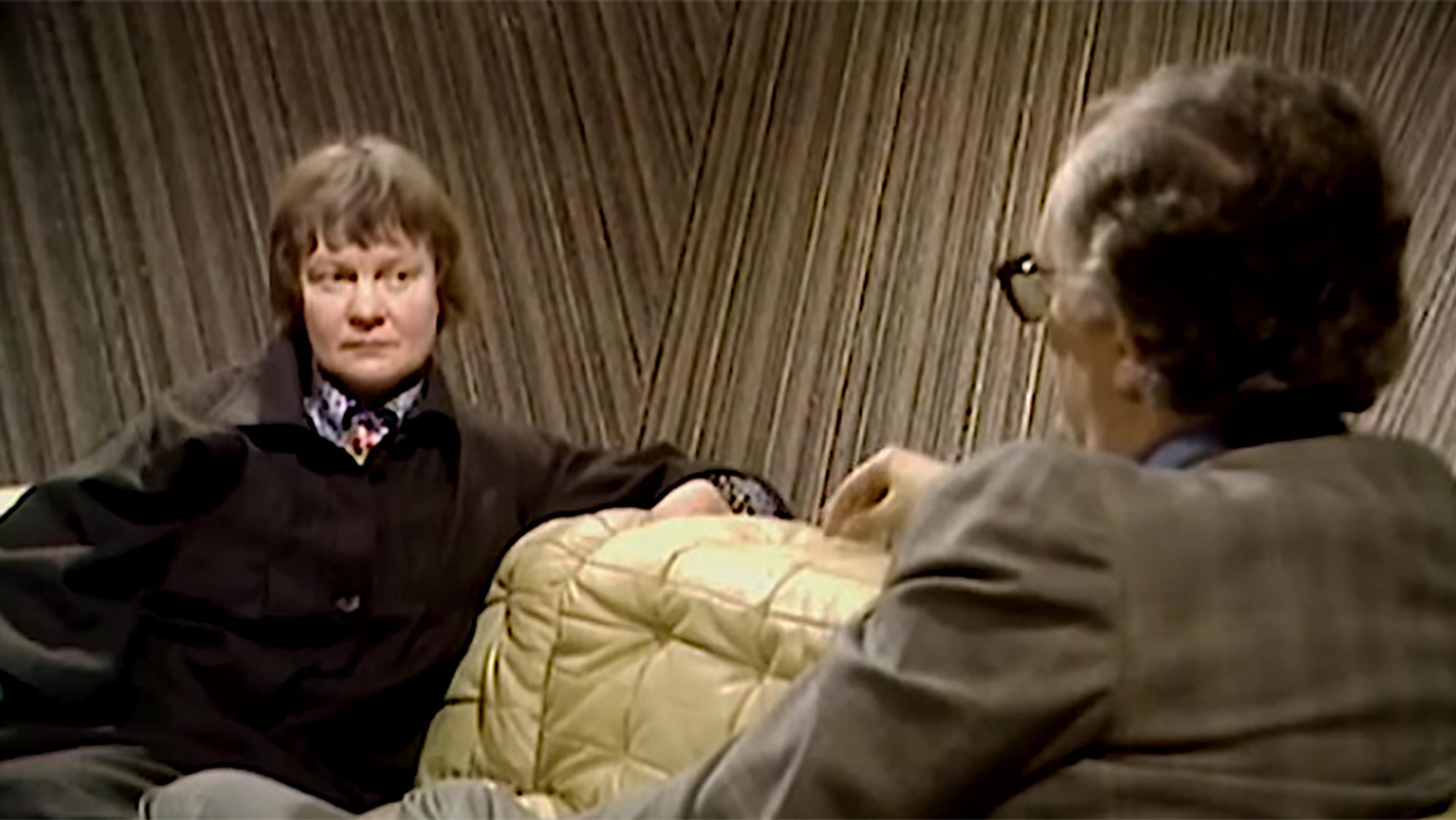 Two people seated and talking on a beige couch, the woman has a serious, attentive expression, the man is viewed from behind. A wood-panelled wall in the background.