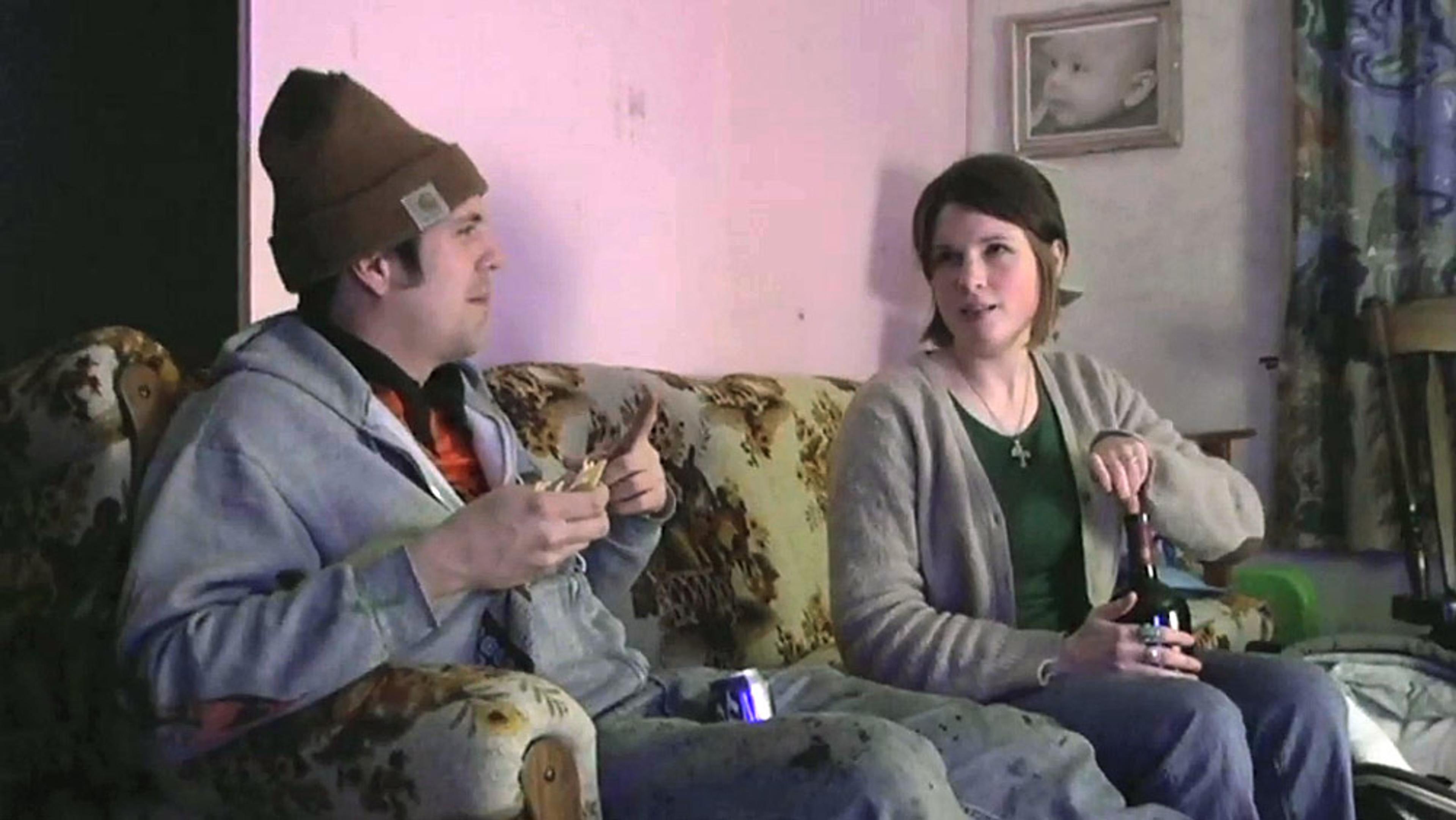 A couple sitting on a patterned sofa, talking. The man holds a sandwich and the woman opens a bottle. A baby photo is on the wall.