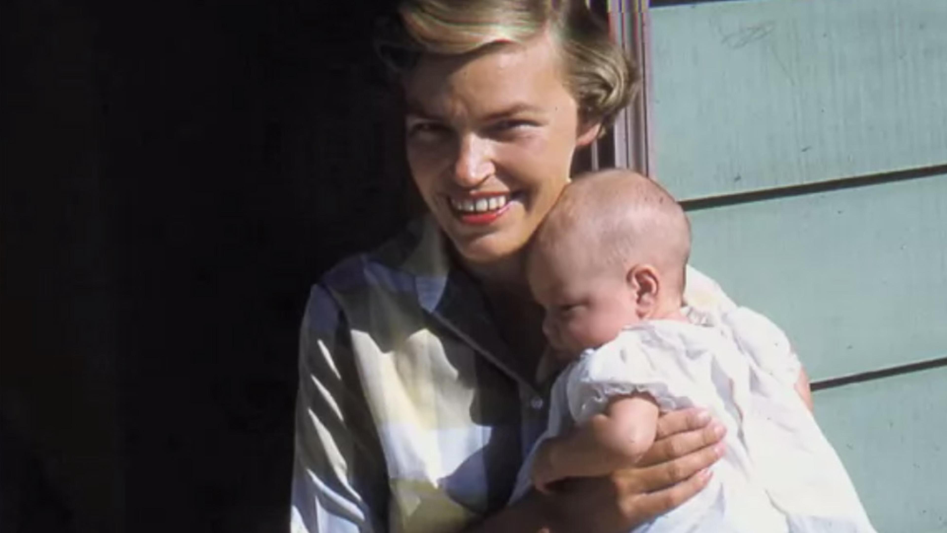 A smiling woman holding a baby in a white dress outside a building with green siding.