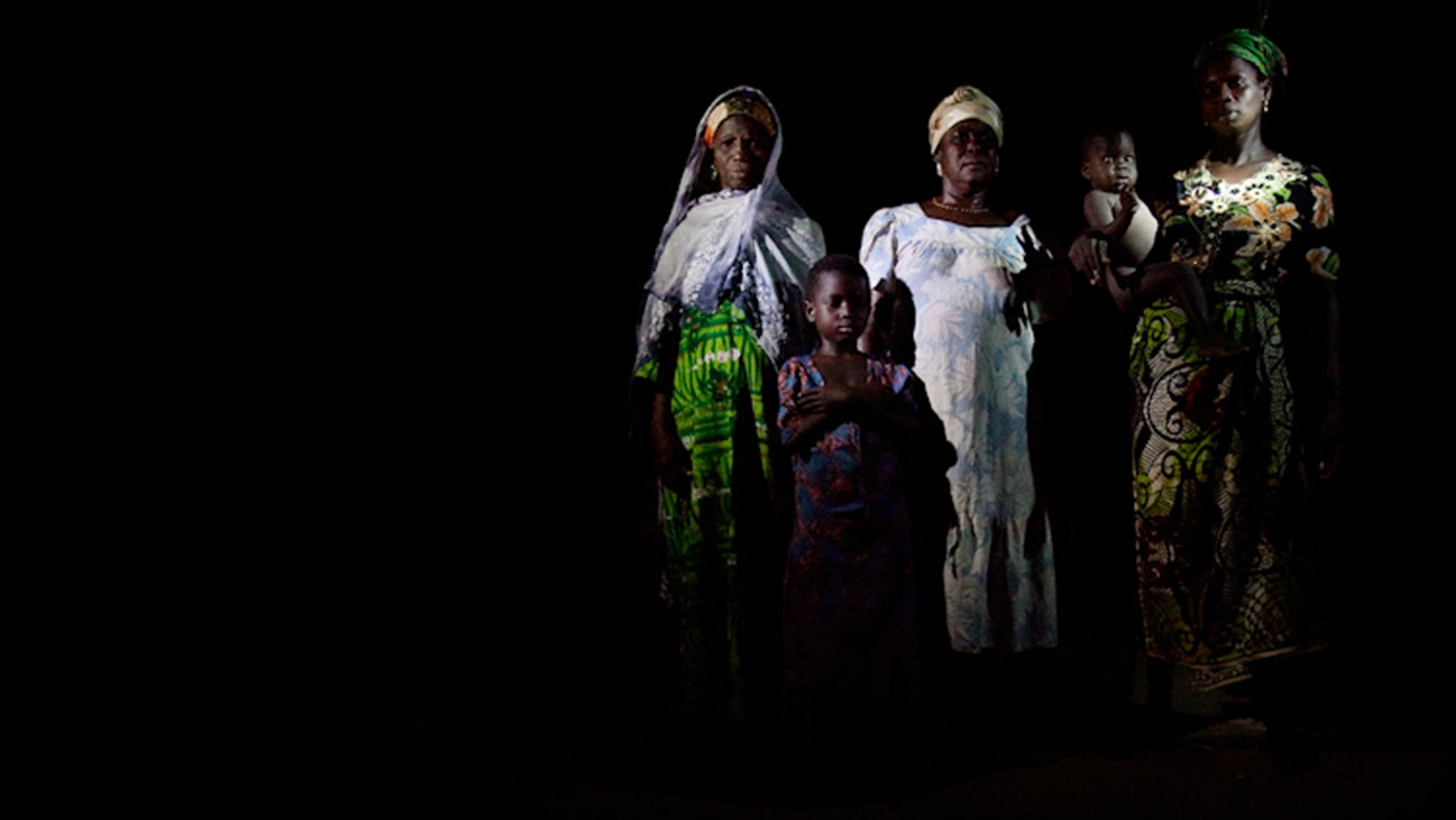 Three women and two children wearing traditional clothing, standing against a dark background.