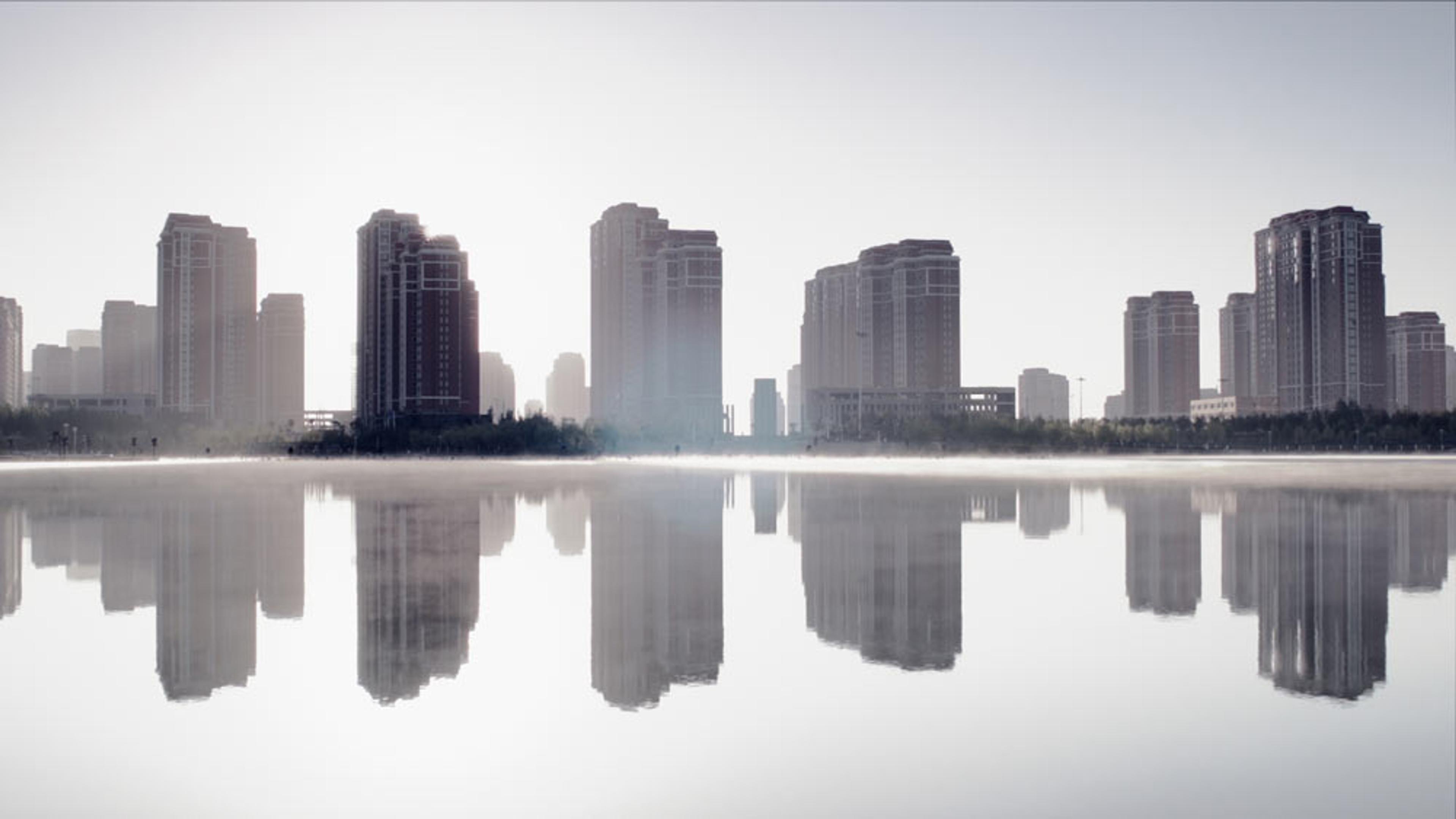 A city skyline with tall buildings reflected in a calm body of water, creating a symmetrical and misty effect.