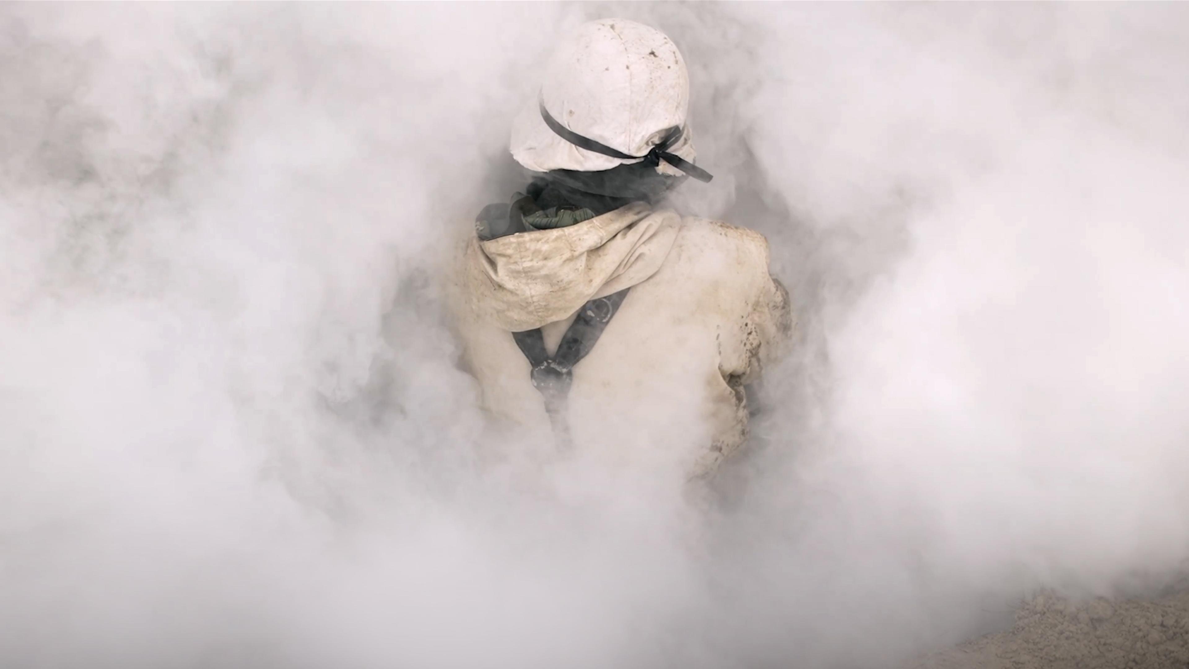 A person in a hard hat and coveralls surrounded by thick white smoke, obscuring most of the view.