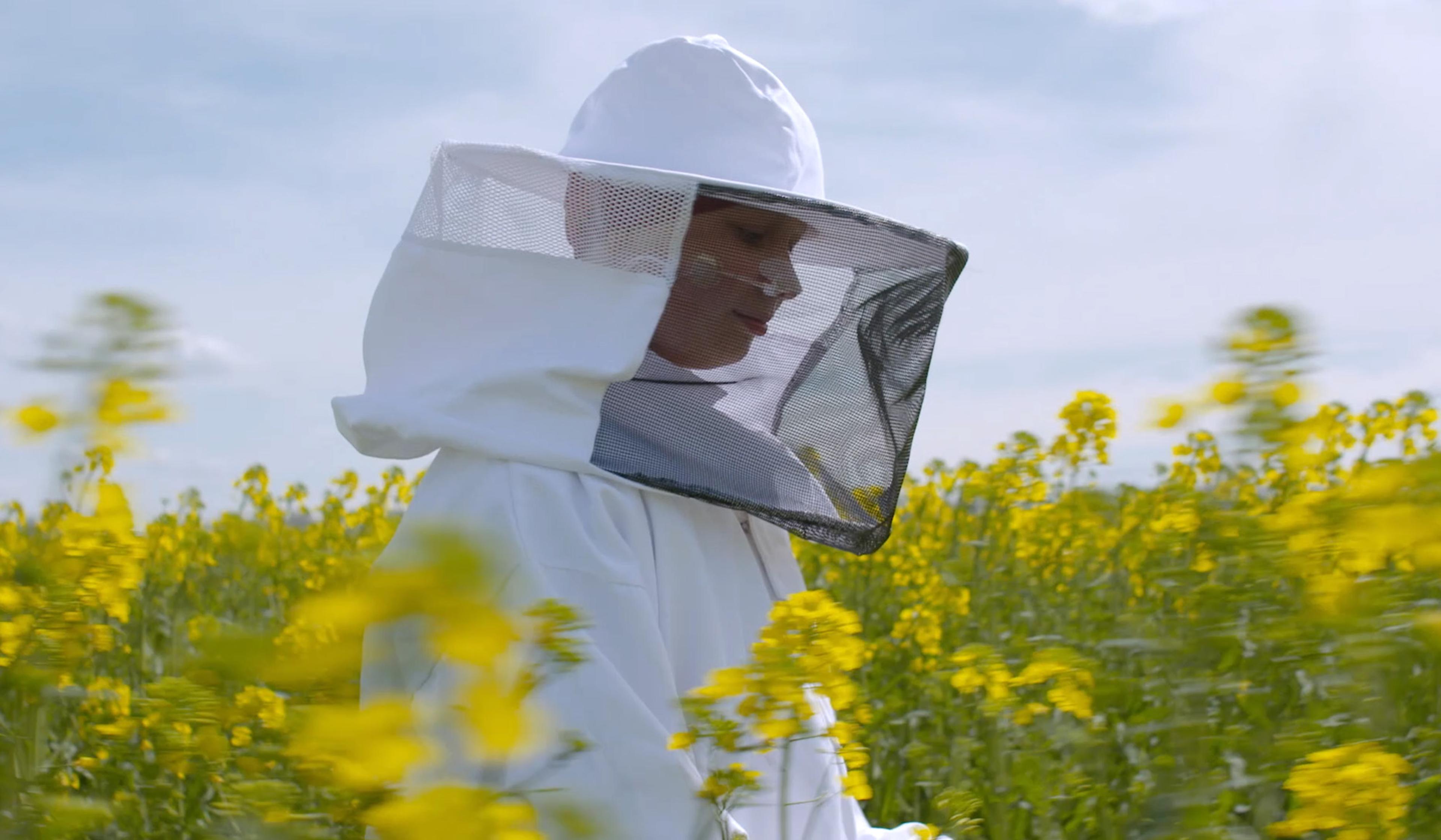 A beekeeper in a white protective suit and veil, standing in a field of yellow flowers under a partly cloudy sky.