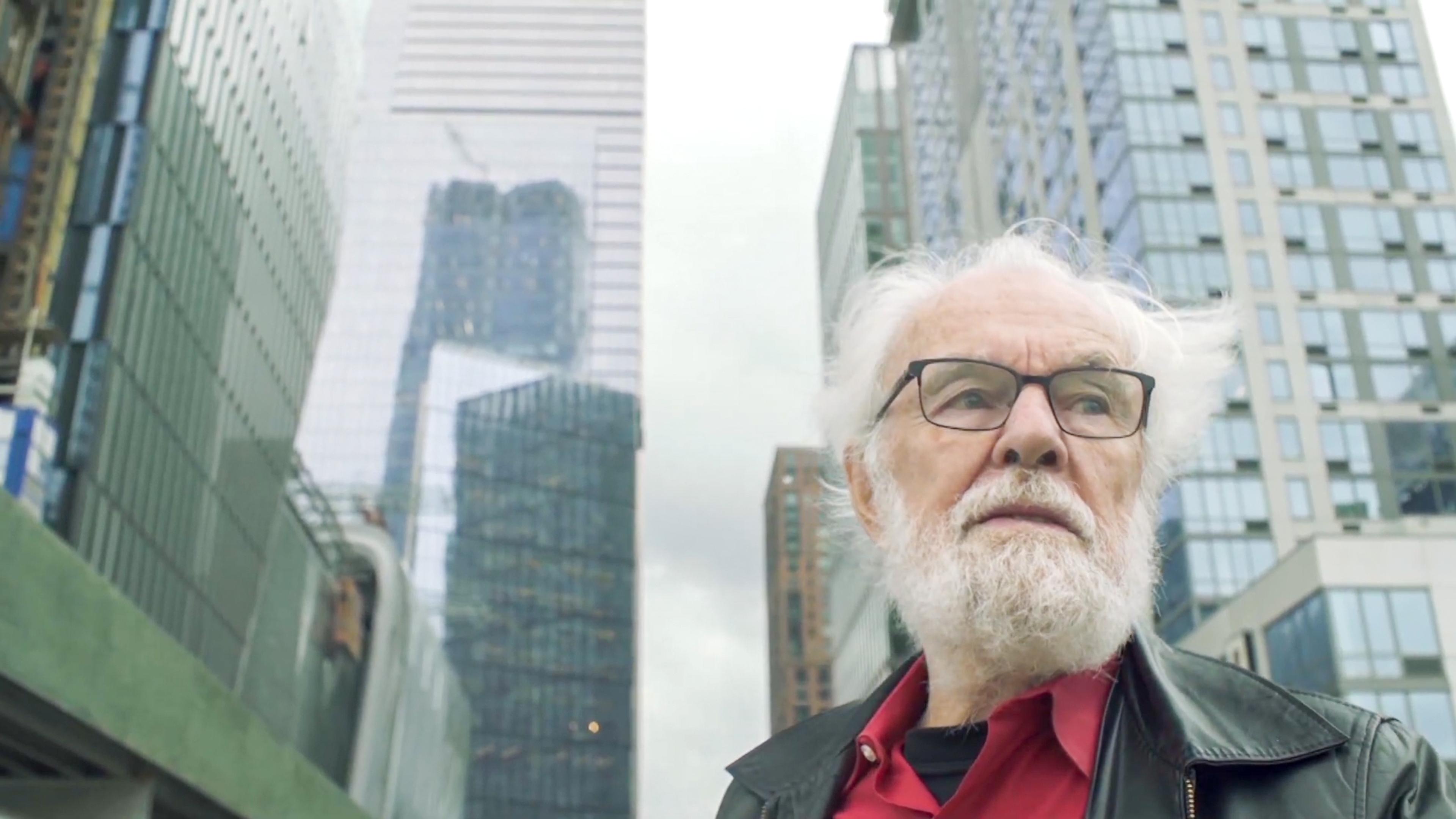Photo of an older man with white hair and beard in a cityscape with tall modern buildings in the background.