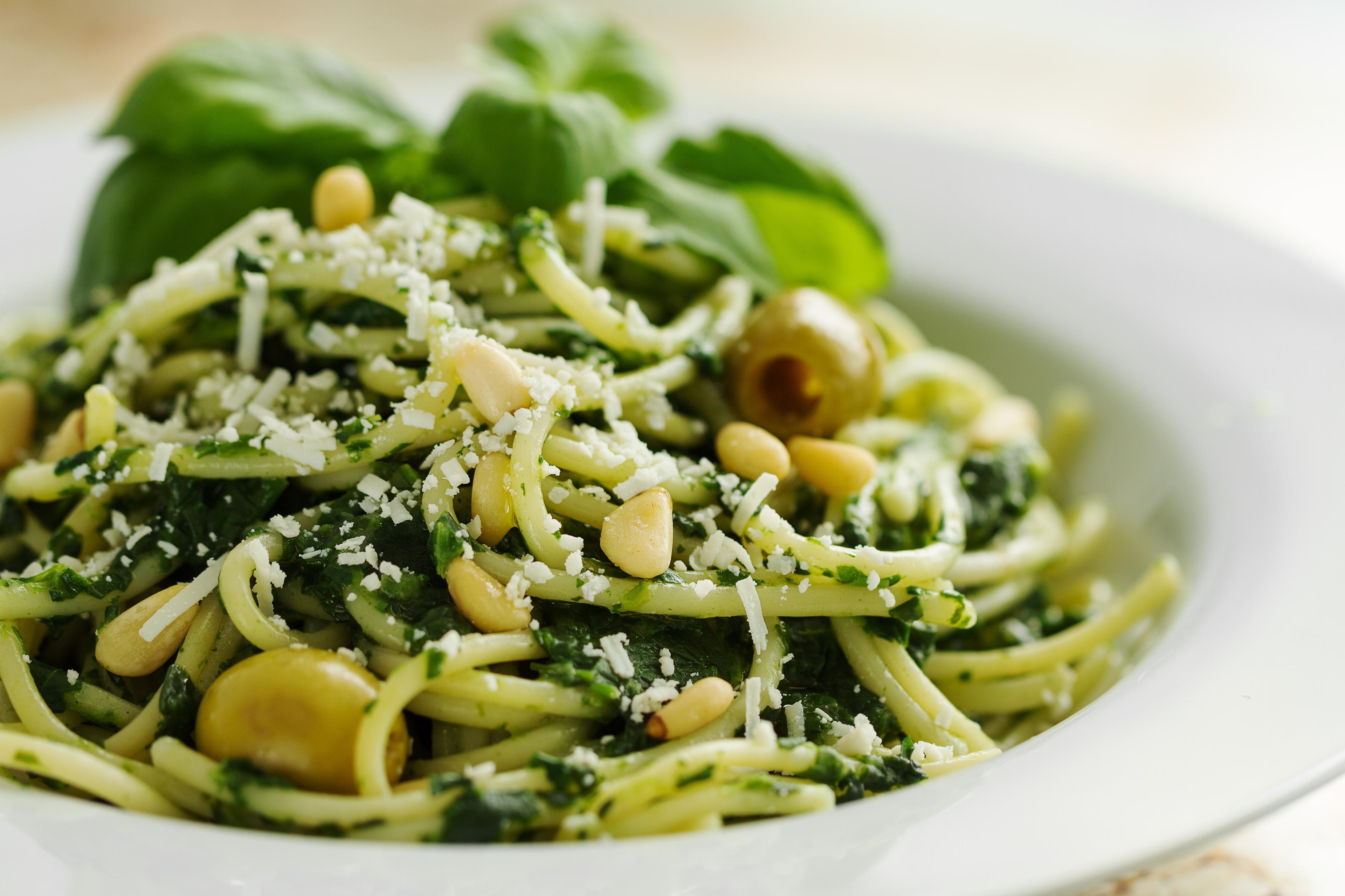 Photo of spaghetti with green sauce, olives and pine nuts garnished with cheese and basil leaves on a white plate.