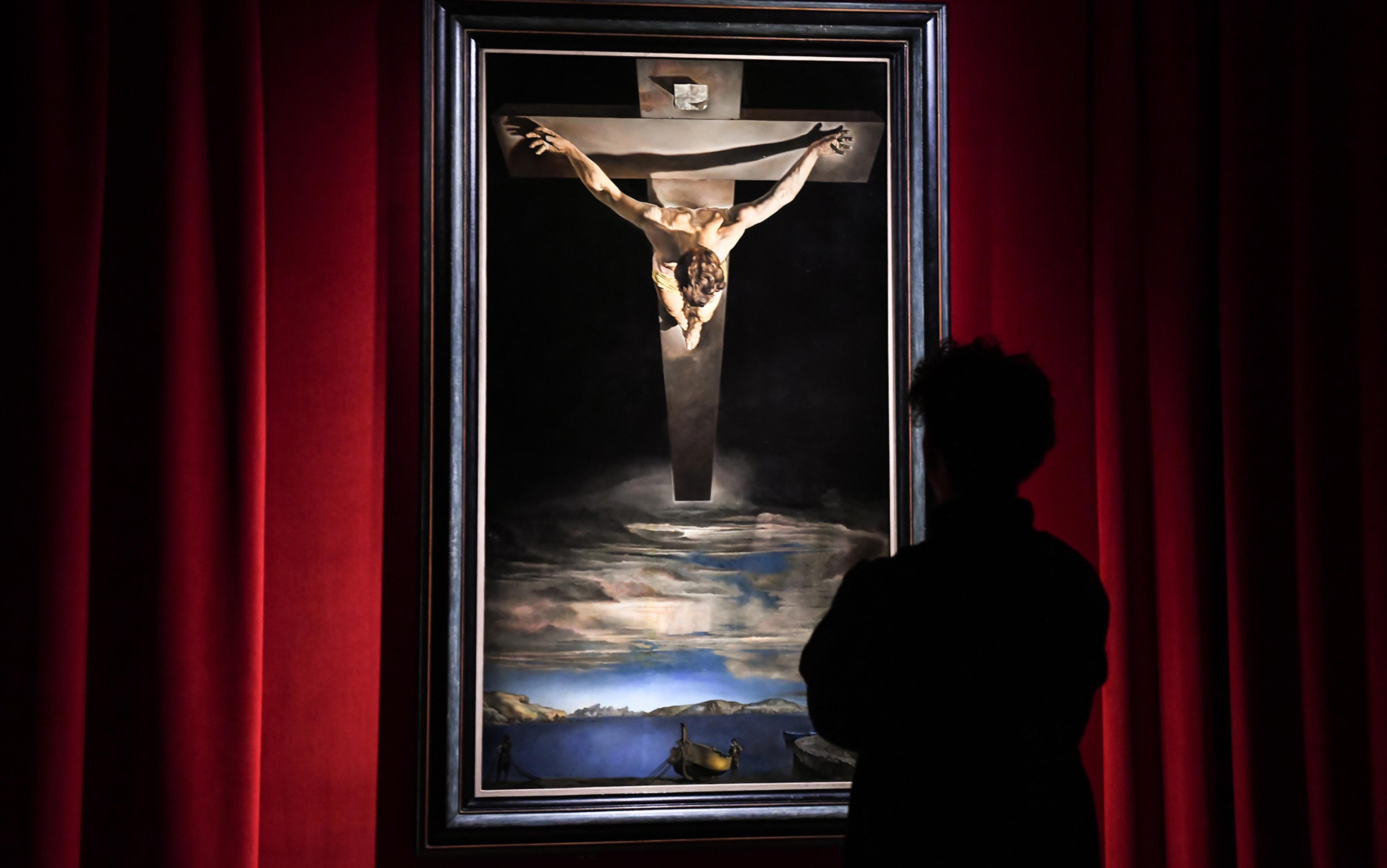 Silhouette of a person viewing a painting of Jesus on the Cross descending into dramatic sky and landscape, framed by red curtains.