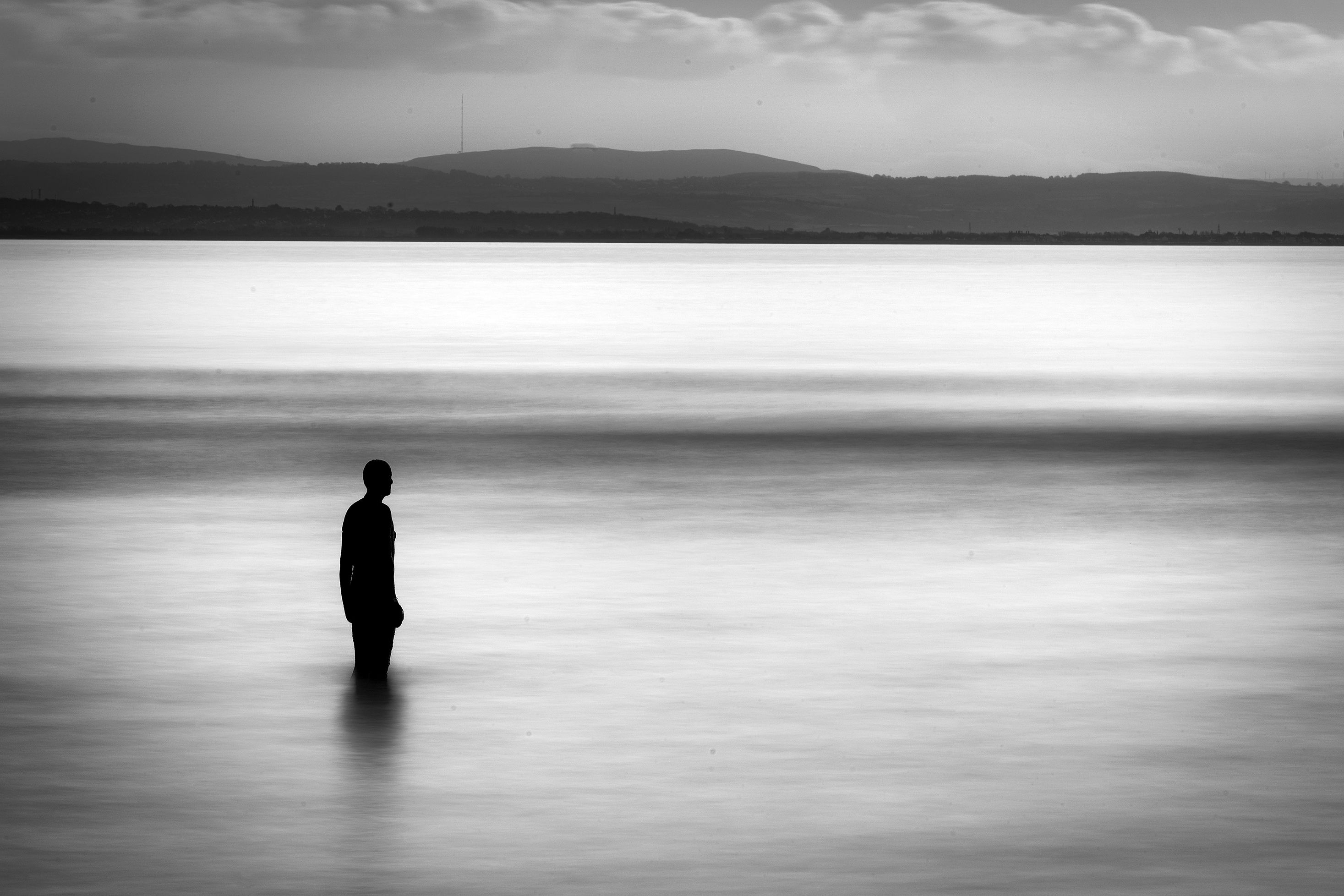 Black and white photo of a silhouette standing in calm water with distant hills and cloudy sky in the background.