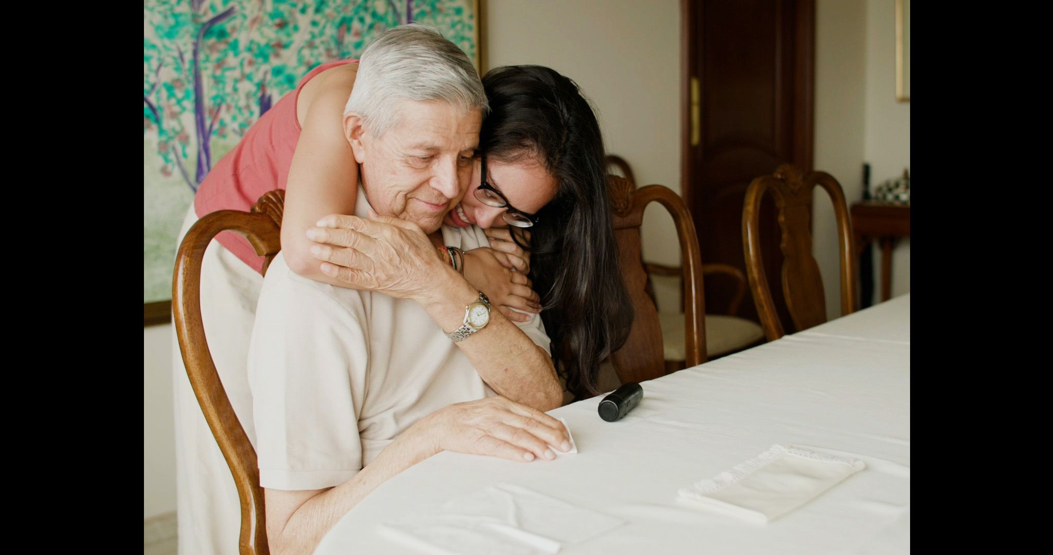 A woman with long dark hair hugs an elderly man sitting at a dining table from behind as they both smile