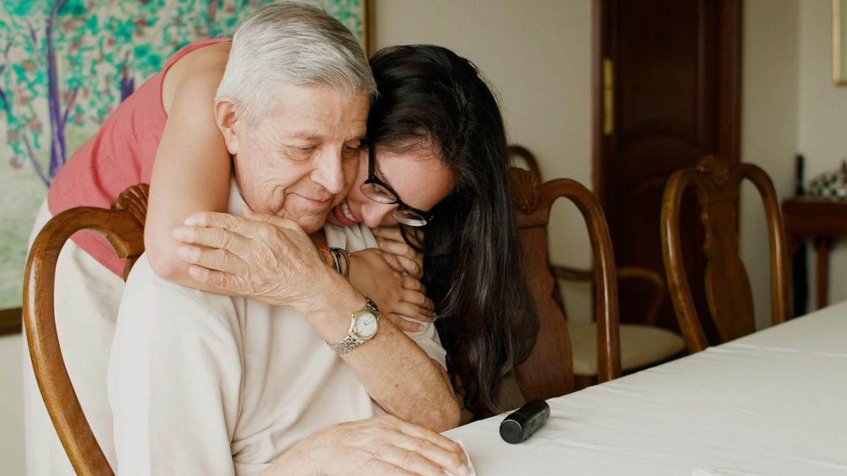 A woman with long dark hair hugs an elderly man sitting at a dining table from behind as they both smile