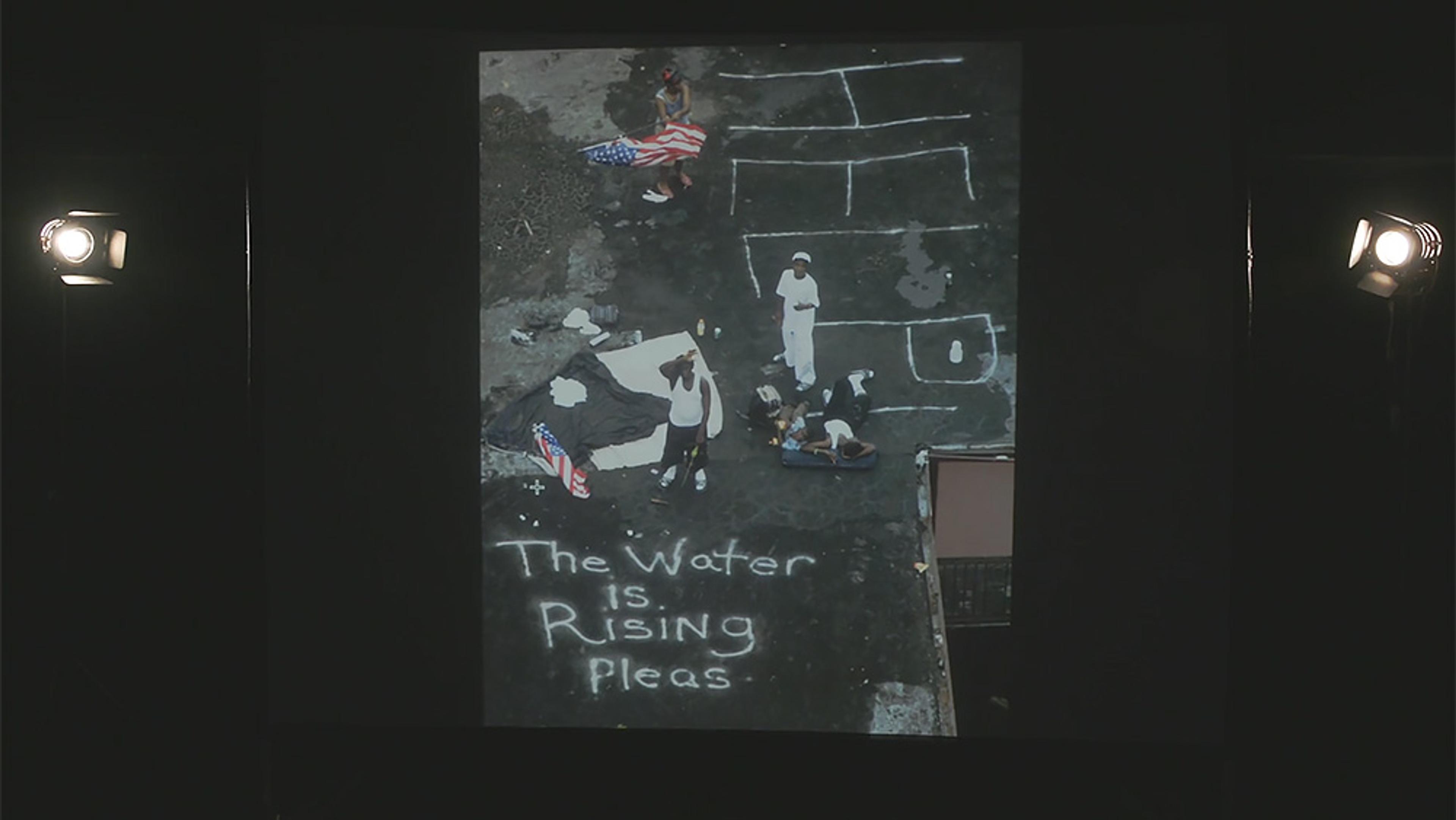 People on a rooftop with “The water is rising pleas” written in chalk. Some hold American flags. Illuminated by two lights.