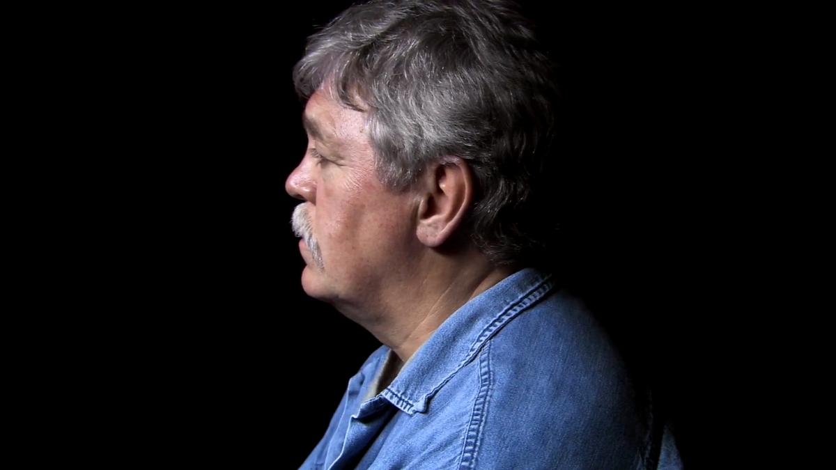 Photo of a man in profile with grey hair and a moustache wearing a denim shirt against a black background.