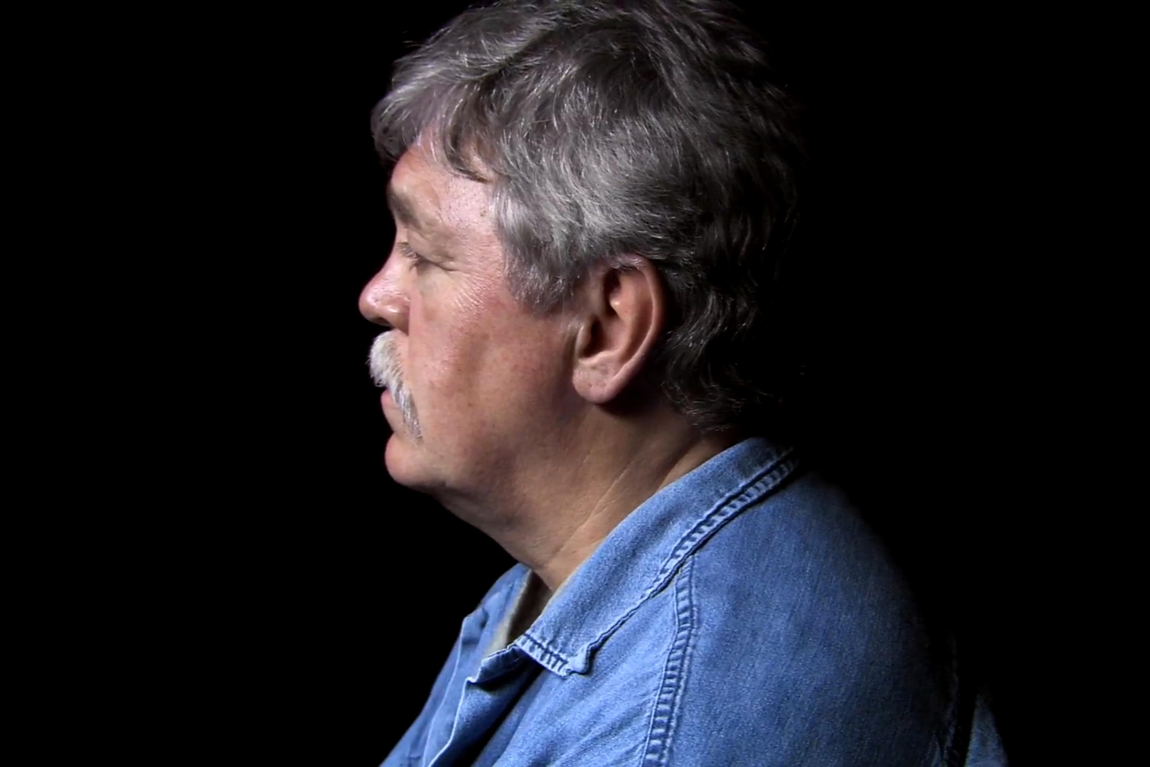 Photo of a man in profile with grey hair and a moustache wearing a denim shirt against a black background.
